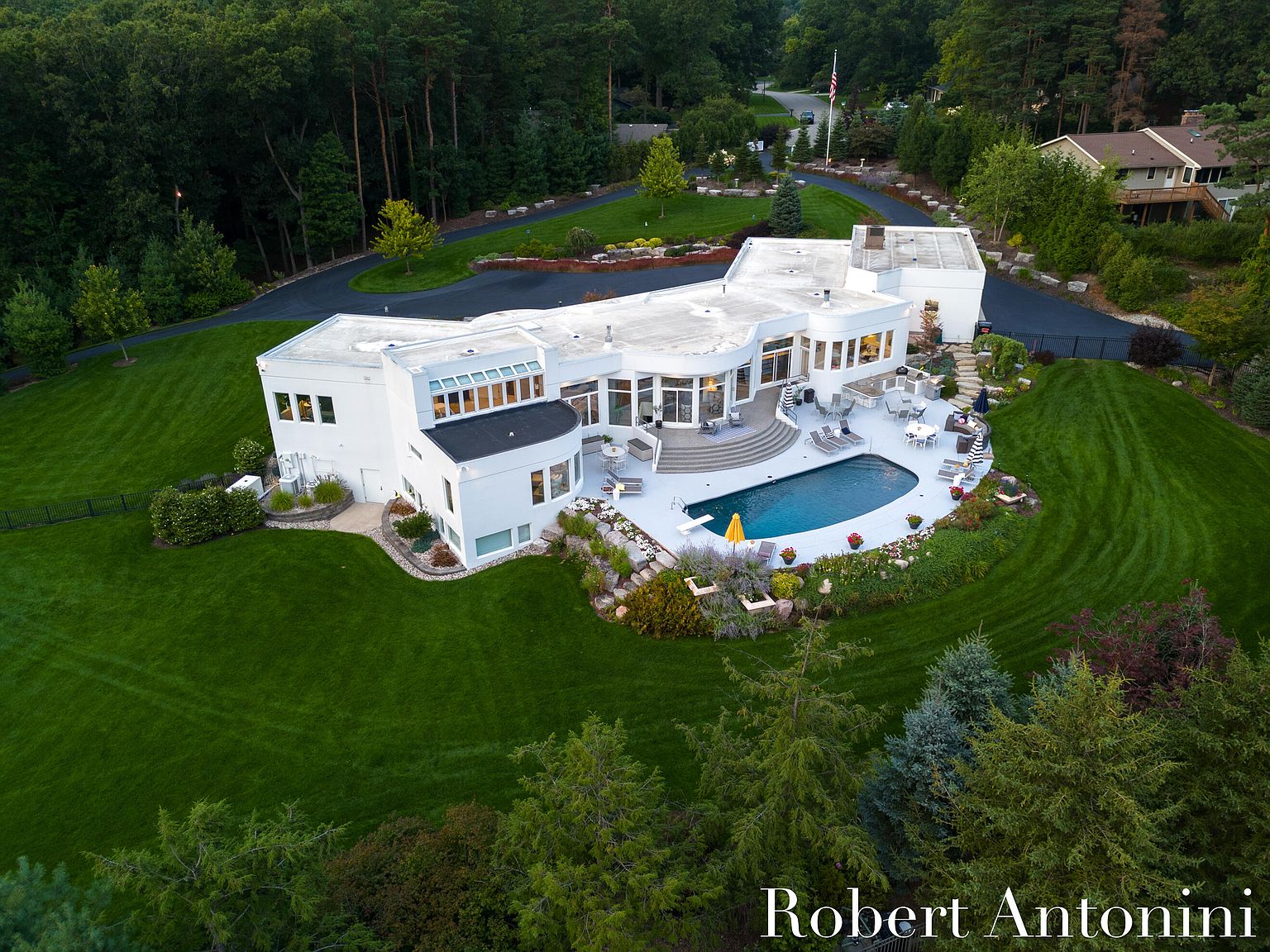 This aerial view showcases a luxurious, modern white home with a unique architectural design. The property features a well-manicured lawn, a private pool with a diving board, and a spacious patio area perfect for entertaining. A long, winding driveway leads to the house, surrounded by lush greenery and mature trees, creating a sense of privacy and exclusivity.