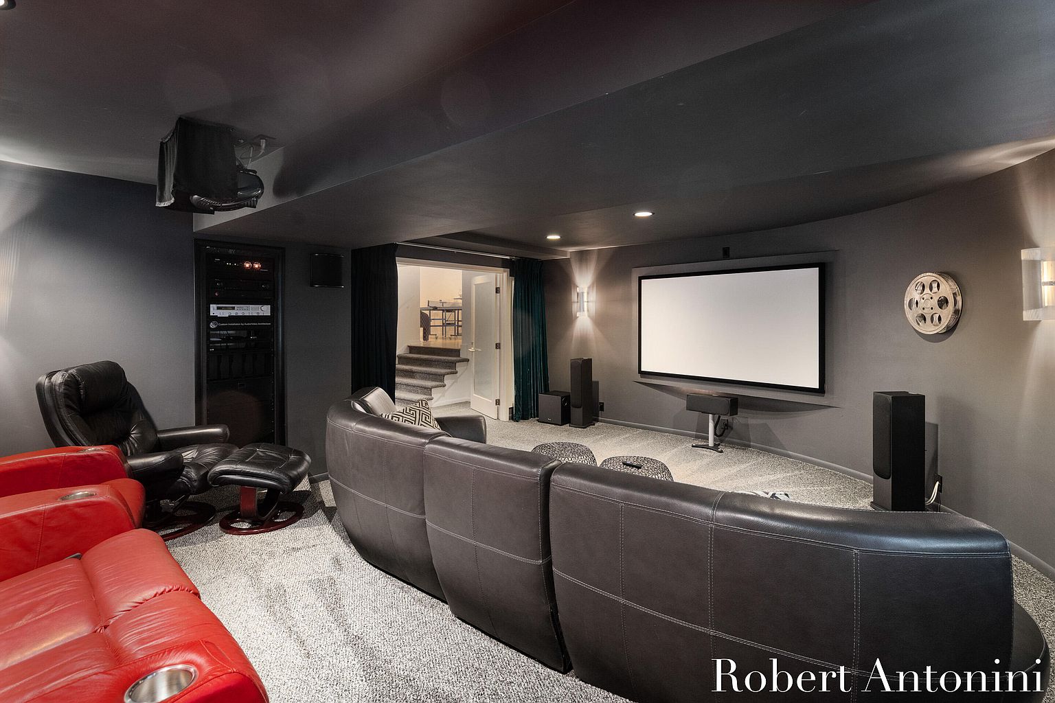 This is a home theater room featuring a large projection screen, comfortable seating including red and black leather recliners, and a sophisticated sound system. The room is painted in dark, neutral tones with a dark ceiling, creating an immersive cinematic experience. A doorway leads to a staircase, and the overall impression is one of luxury and entertainment.
