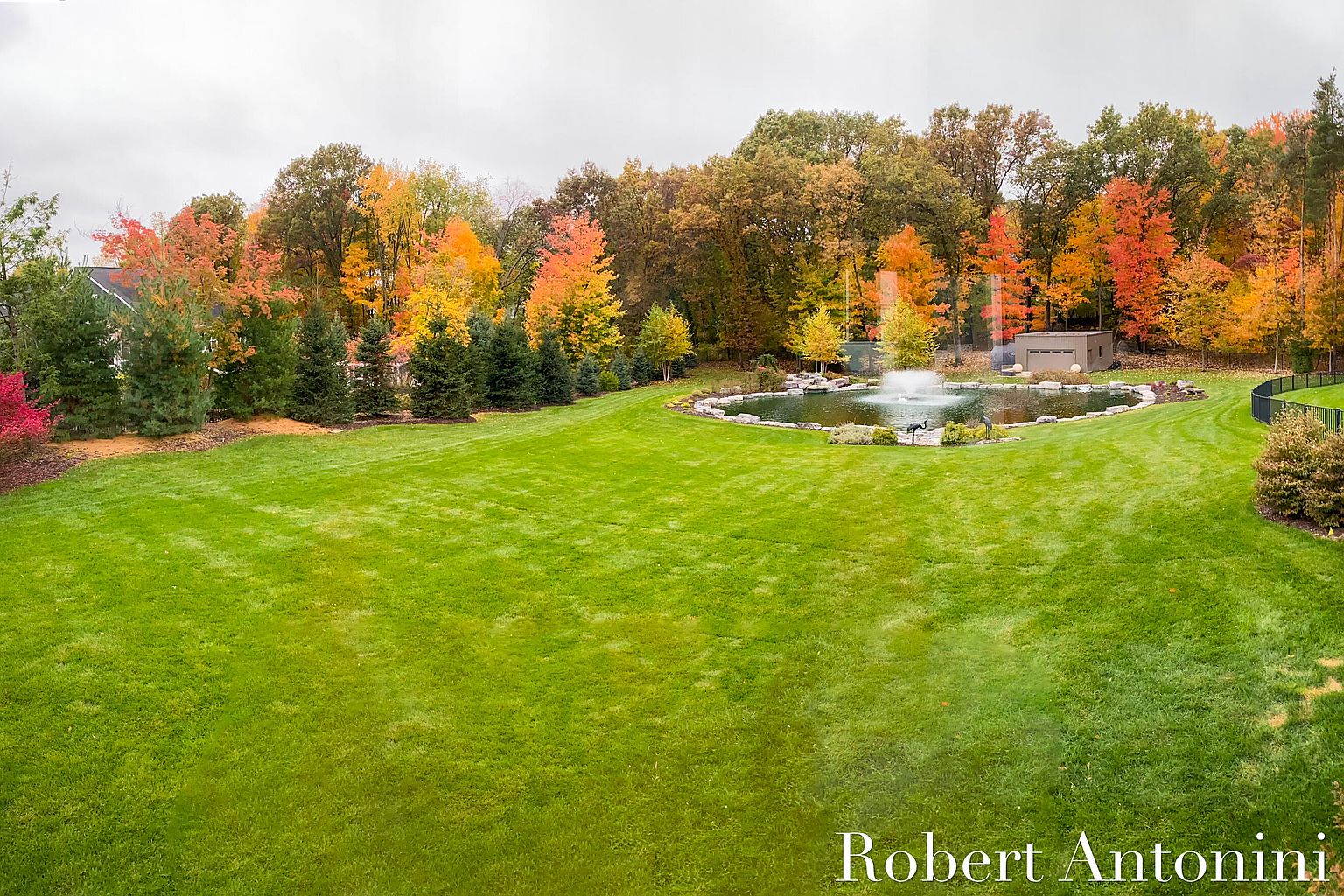 This image showcases a meticulously maintained backyard featuring a lush green lawn, a serene pond with a fountain, and vibrant autumn foliage. The landscape is well-manicured, creating a picturesque and inviting outdoor space. A small outbuilding is visible near the pond, adding to the property's appeal.