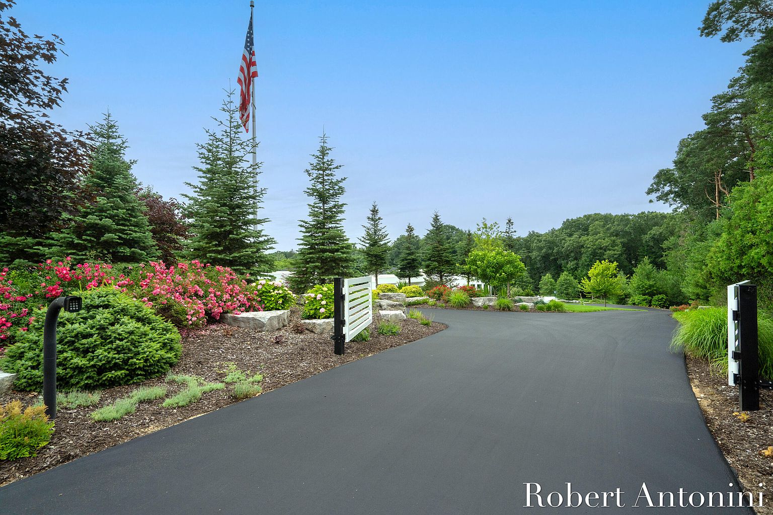 This image showcases a beautifully landscaped yard and driveway leading up to a property. The scene features lush greenery, including various trees, bushes, and flowering plants, complemented by decorative rocks and mulch. An American flag stands tall in the background, adding a patriotic touch to the well-maintained exterior.