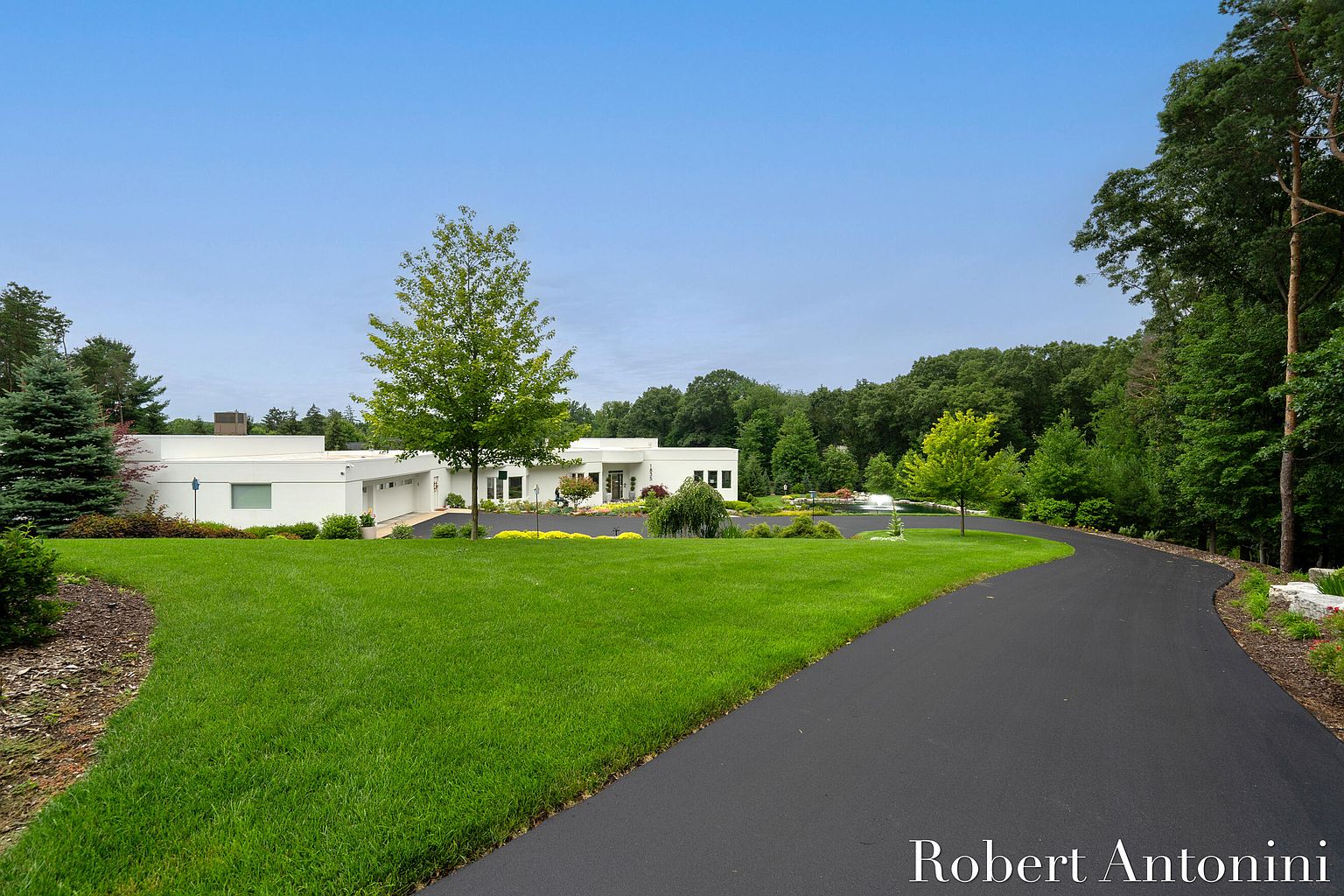 This image showcases the front exterior of a modern, single-story home with a sprawling green lawn and a curved driveway. The house features a clean, white facade with minimalist architectural details, complemented by mature trees and landscaping. The overall impression is one of understated elegance and serene privacy.