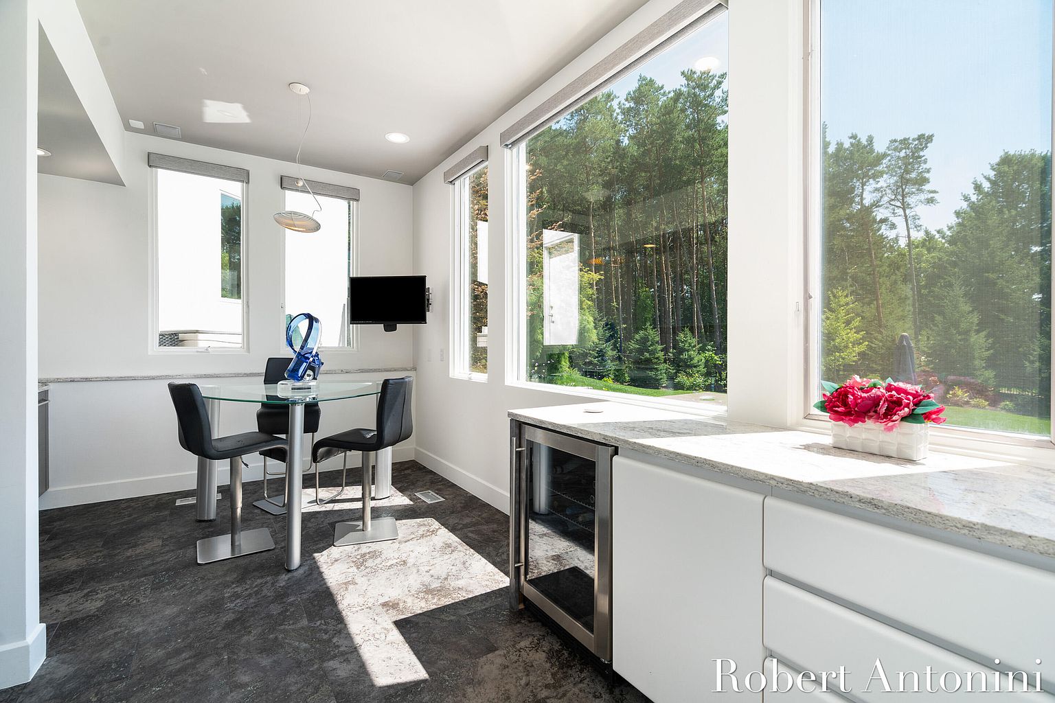 This is a bright and modern dining area featuring a glass-top table with four black chairs. Large windows offer a view of the lush greenery outside, enhancing the natural light in the room. A built-in wine cooler and sleek white cabinetry add to the contemporary aesthetic.