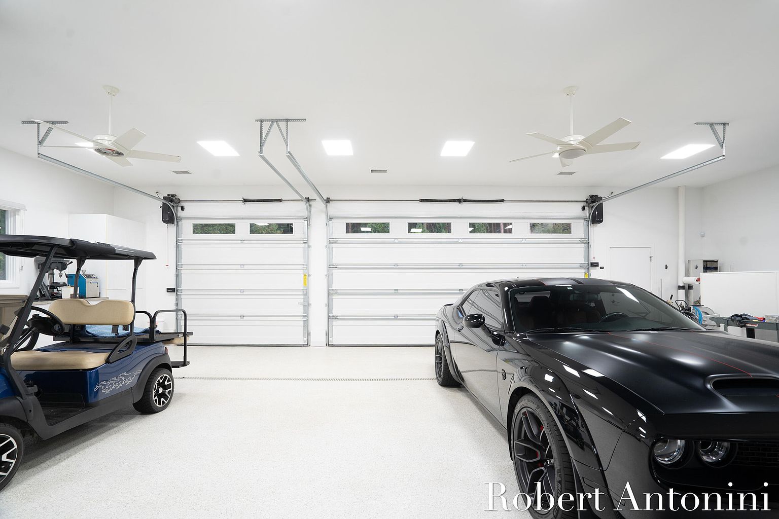 This is a well-organized garage featuring a black car and a golf cart. The garage has two white garage doors and a white ceiling with ceiling fans and recessed lighting. The floor is a speckled white epoxy, and the overall impression is clean and modern.