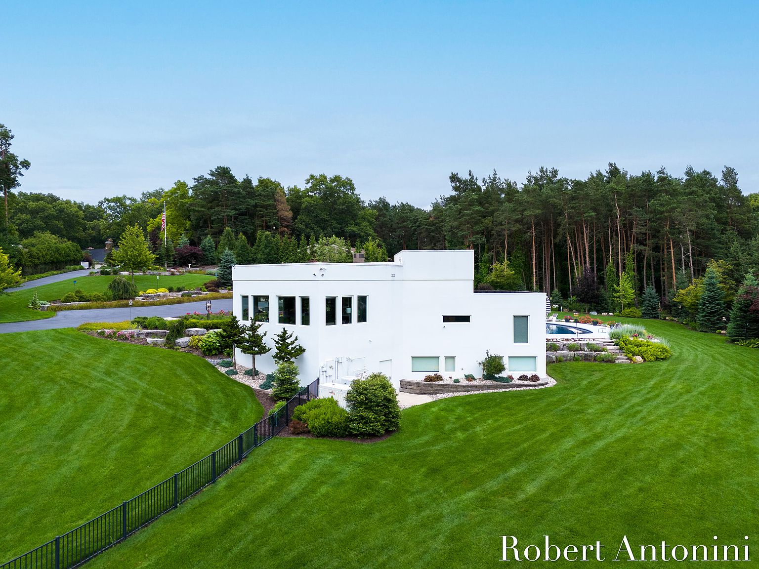This aerial view showcases a modern, white home surrounded by lush green lawns and mature trees. The property features a swimming pool and well-maintained landscaping, creating a serene and private setting. A black fence borders the property, adding a touch of elegance.