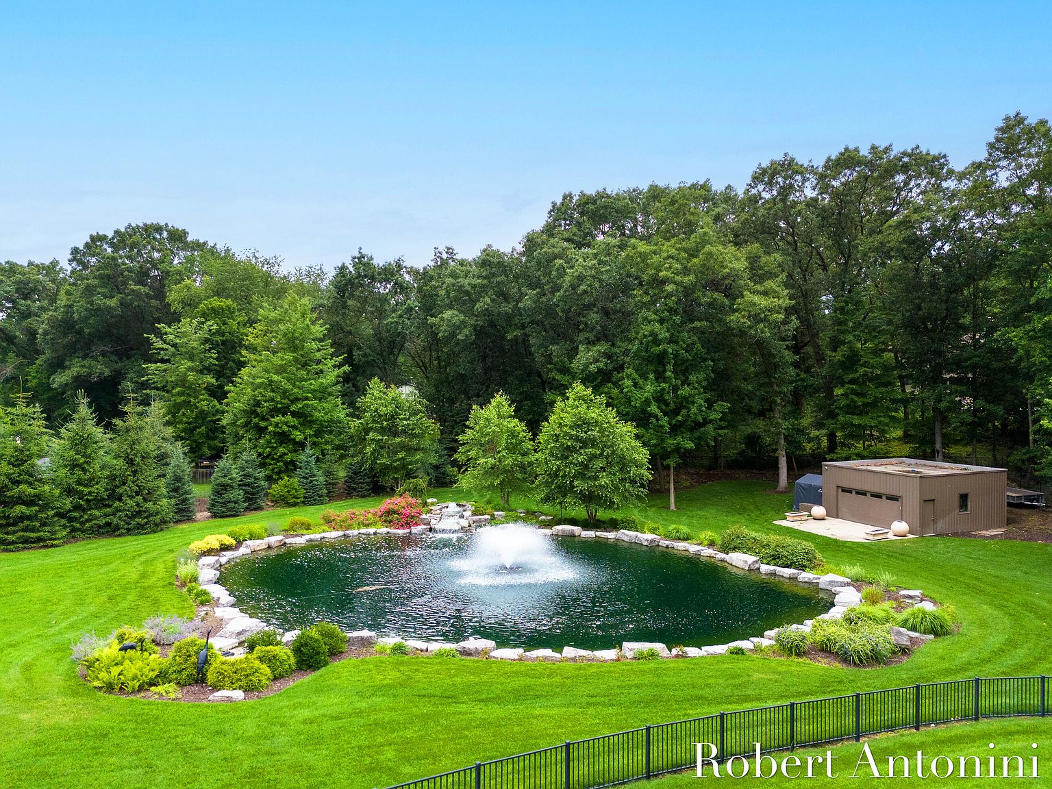 This aerial view showcases a meticulously landscaped yard featuring a large, oval-shaped pond with a central fountain. The pond is surrounded by rocks and lush greenery, including various plants and trees. A modern outbuilding is visible in the background, adding a contemporary touch to the natural setting. A black fence is visible in the foreground.