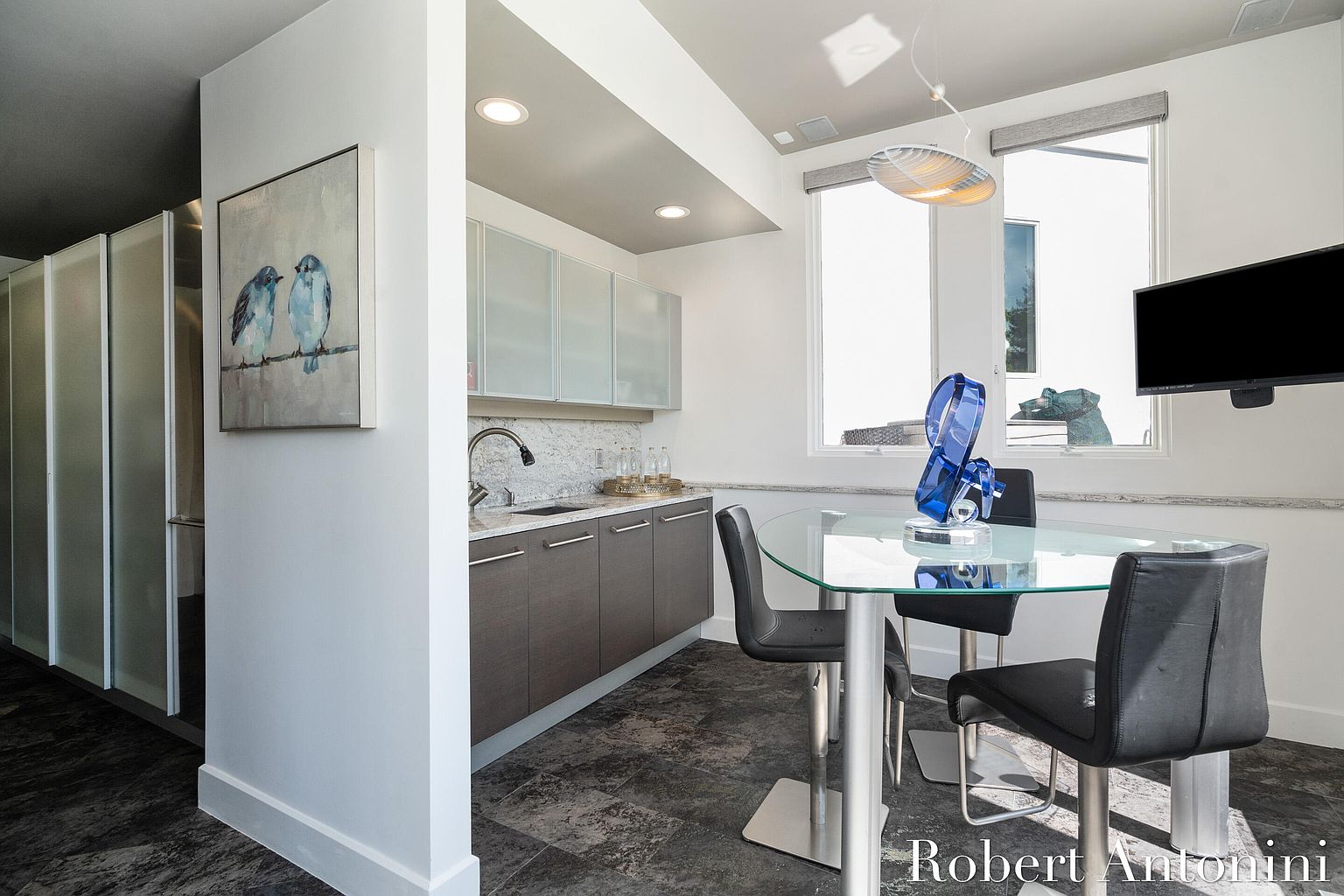 This interior shot showcases a modern dining area with a glass-top table surrounded by sleek black chairs. A built-in bar area with gray cabinets and a marble backsplash adds functionality and style. Natural light streams in through the windows, highlighting the contemporary design and dark tile flooring.