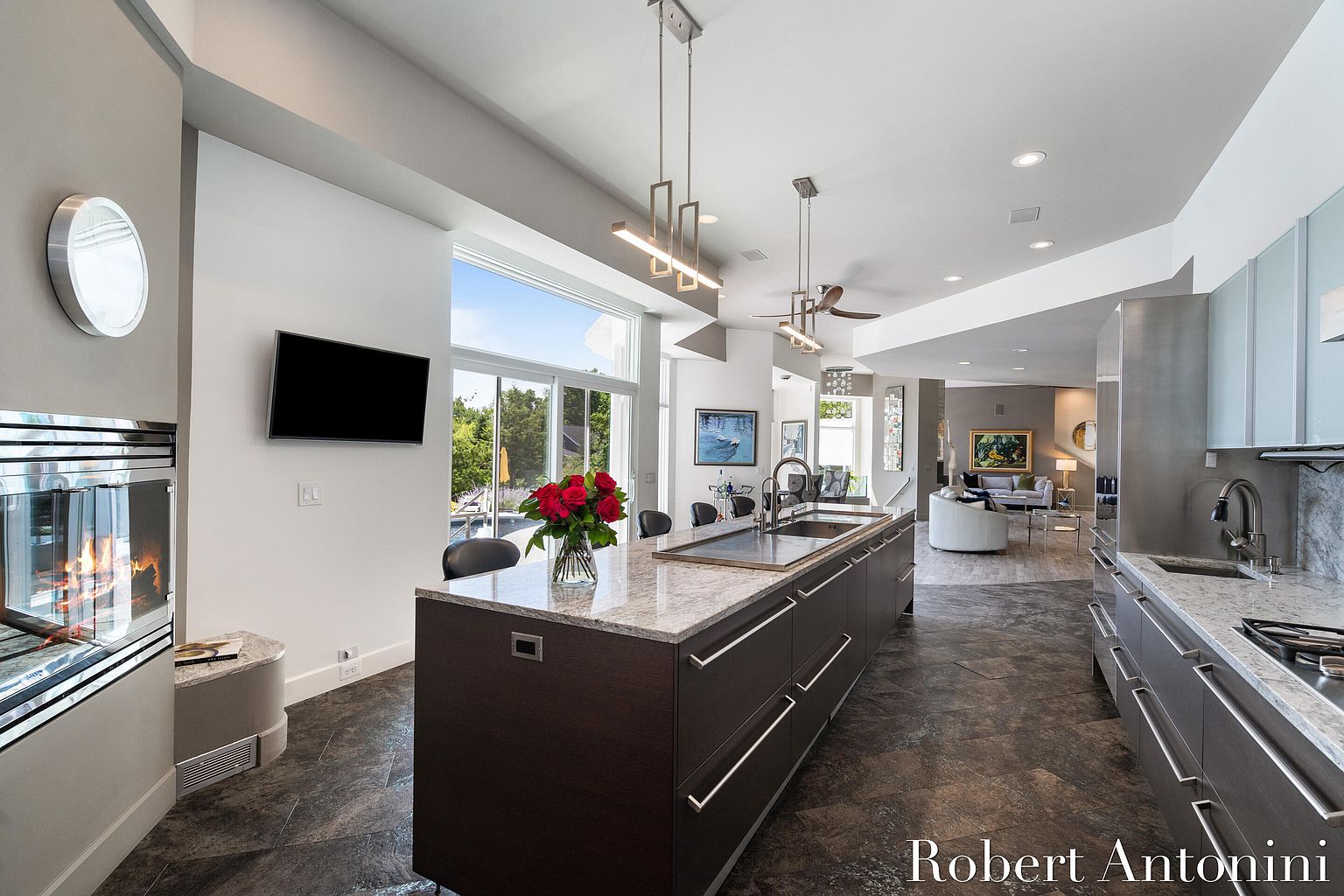 This is a modern kitchen featuring a large island with a granite countertop and dark wood cabinetry. The kitchen has stainless steel appliances and a view of the outdoor area through large windows. The flooring is a dark tile, and the space is illuminated by modern pendant lighting.
