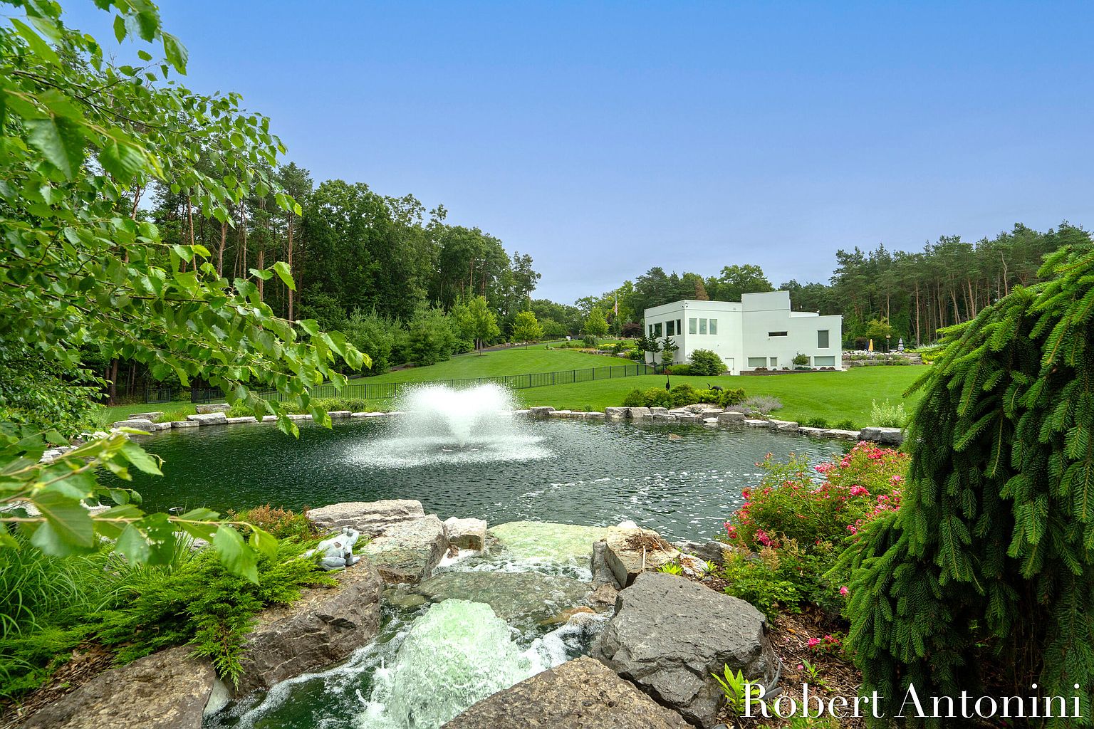 This exterior shot showcases a beautifully landscaped yard featuring a serene pond with a fountain and a waterfall feature. The modern white house is visible in the background, surrounded by lush green lawns and mature trees. The scene evokes a sense of tranquility and luxury, highlighting the property's expansive outdoor space and meticulous landscaping.