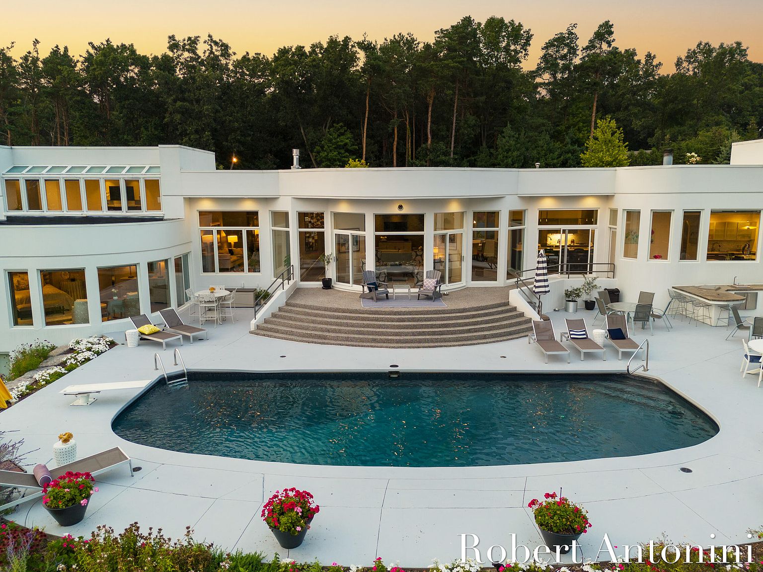 This aerial view showcases a luxurious backyard featuring a modern white house with large windows and a spacious patio. A sleek, dark-water pool is the focal point, surrounded by lounge chairs, potted flowers, and well-maintained landscaping. The scene evokes a sense of relaxation and upscale outdoor living.