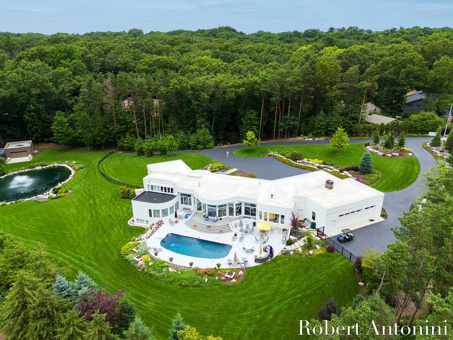 This aerial view showcases a luxurious modern home with a sprawling green lawn, a large swimming pool, and a private pond. The house features a white exterior, a curved architectural design, and an attached garage. The property is surrounded by lush trees, providing a sense of privacy and tranquility.