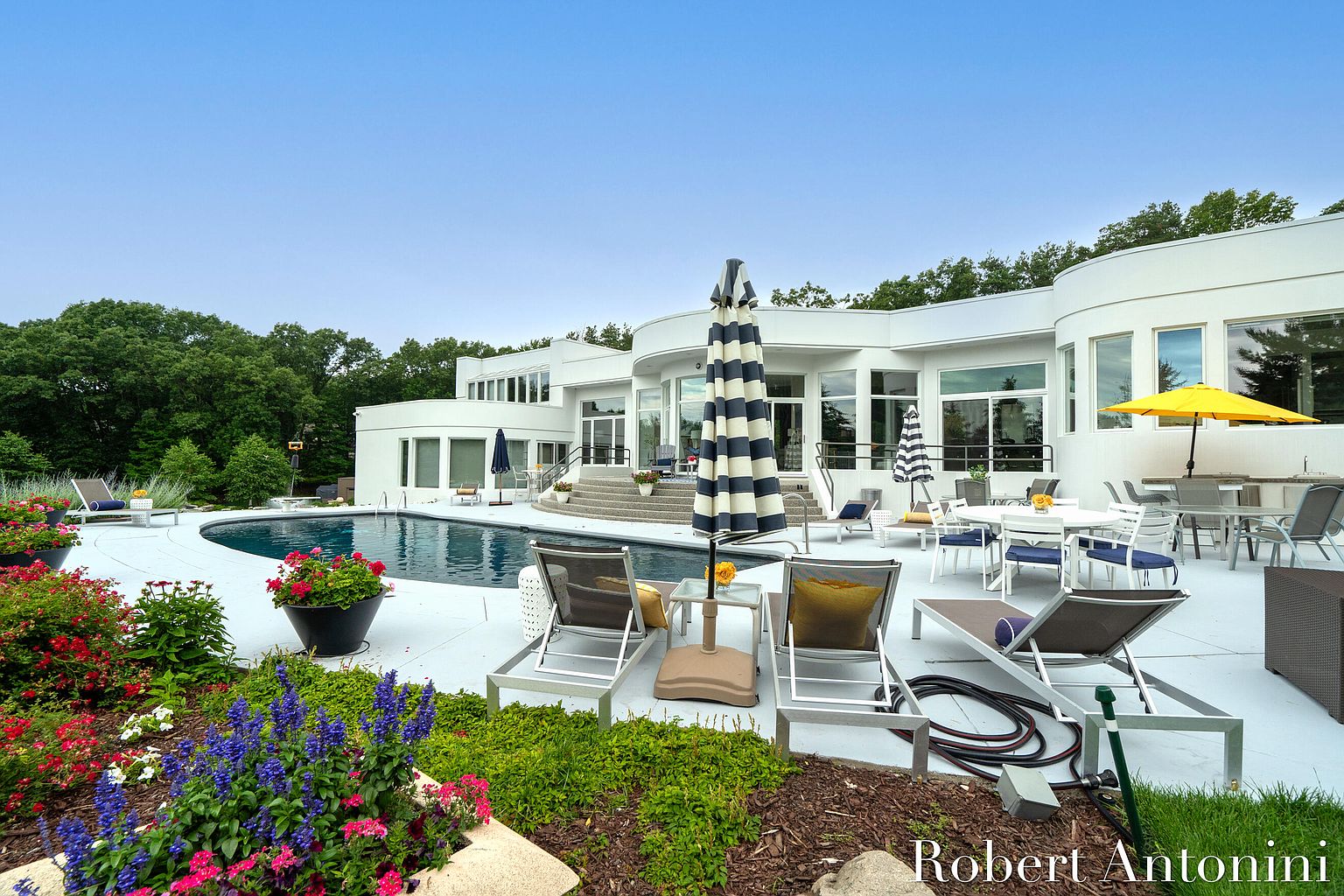 This image showcases a luxurious backyard with a modern, white-exterior home as the backdrop. A curved swimming pool is the focal point, surrounded by a white patio area furnished with lounge chairs and tables with umbrellas. The landscaping includes colorful flower beds and lush greenery, creating a serene and inviting outdoor living space.