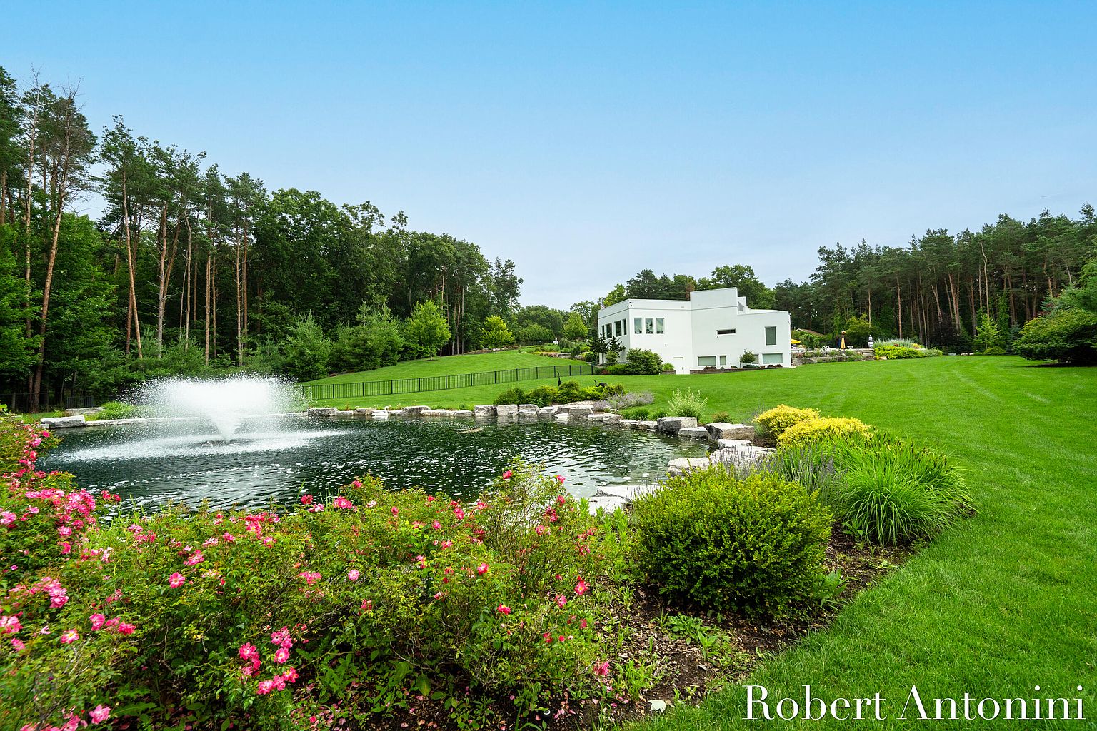This exterior shot showcases a meticulously landscaped yard featuring a serene pond with a fountain as a focal point. The modern white house is nestled in the background, surrounded by lush green lawns and mature trees. The scene exudes tranquility and highlights the property's expansive outdoor space, perfect for relaxation and entertainment.