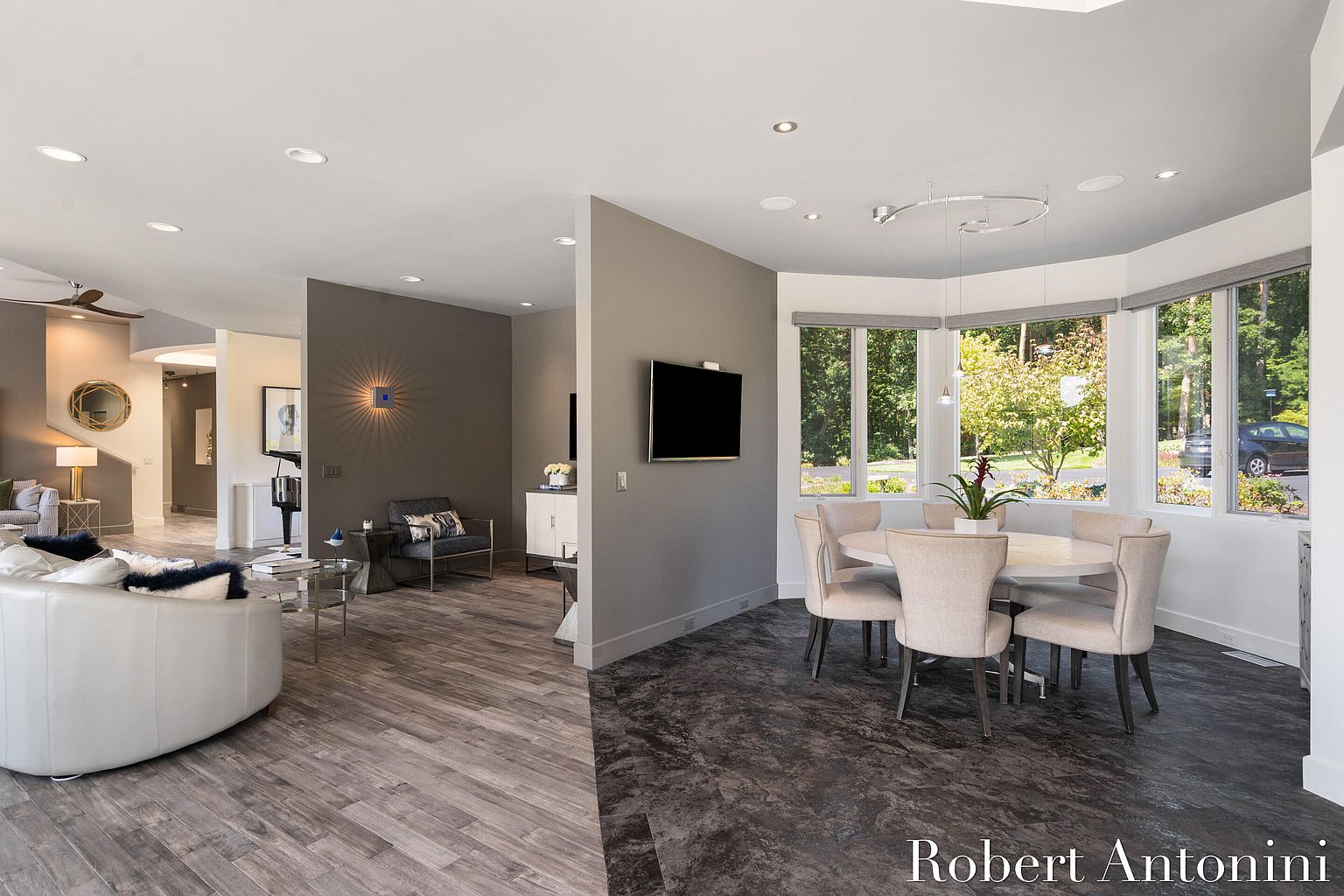 This interior shot showcases a dining area adjacent to a living space, featuring a round dining table with six chairs positioned near a bay window that offers natural light and views of the outdoors. The flooring transitions from wood in the living area to a darker, textured material in the dining space, creating visual separation. The room is decorated in neutral tones, providing a sophisticated and inviting atmosphere.