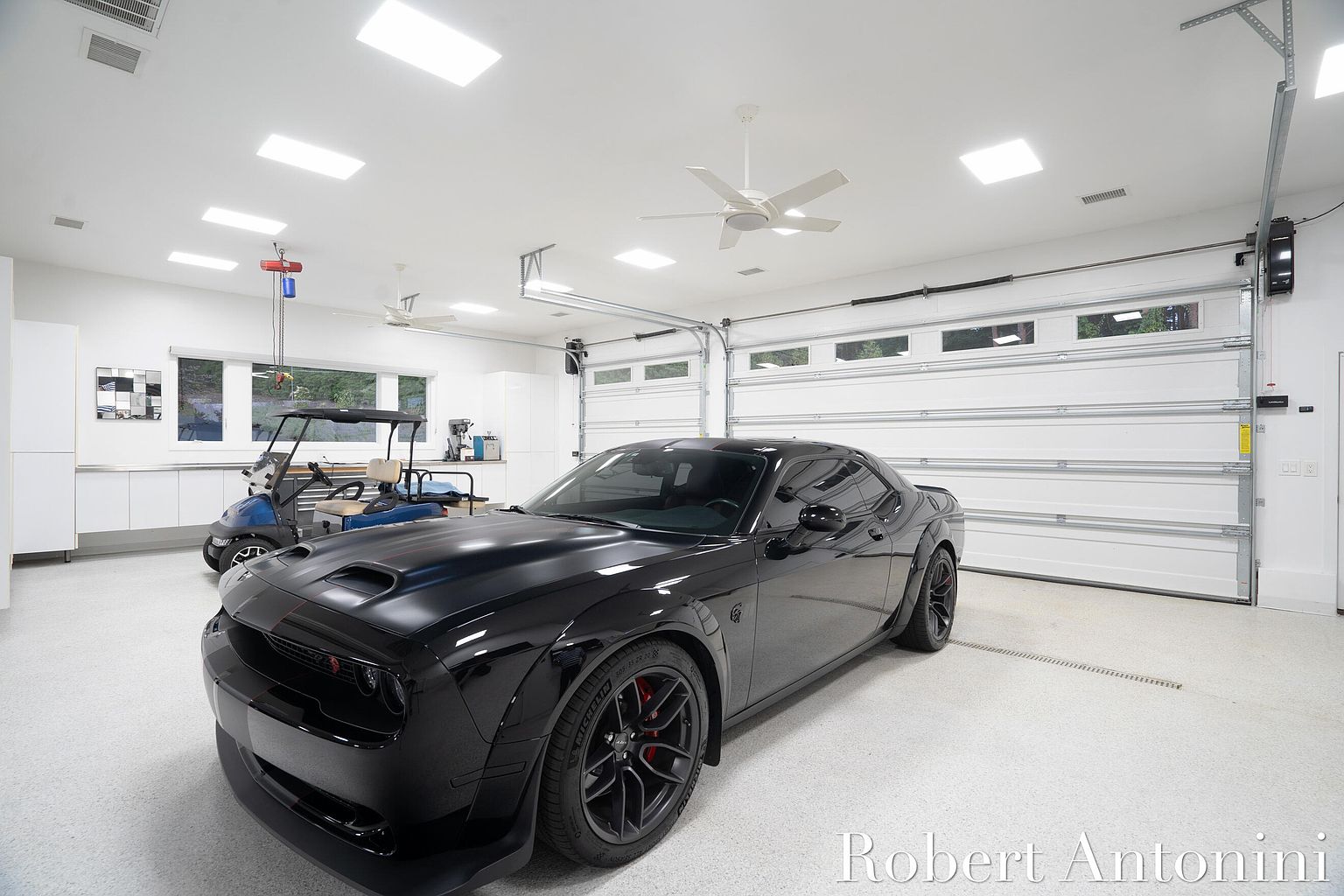 This is a well-lit, modern garage featuring a sleek black car as the focal point. The garage has a clean, white interior with a high ceiling, recessed lighting, and a white epoxy floor. A golf cart is parked in the background, and a large white garage door with windows is visible, contributing to the overall spacious and organized feel of the garage.
