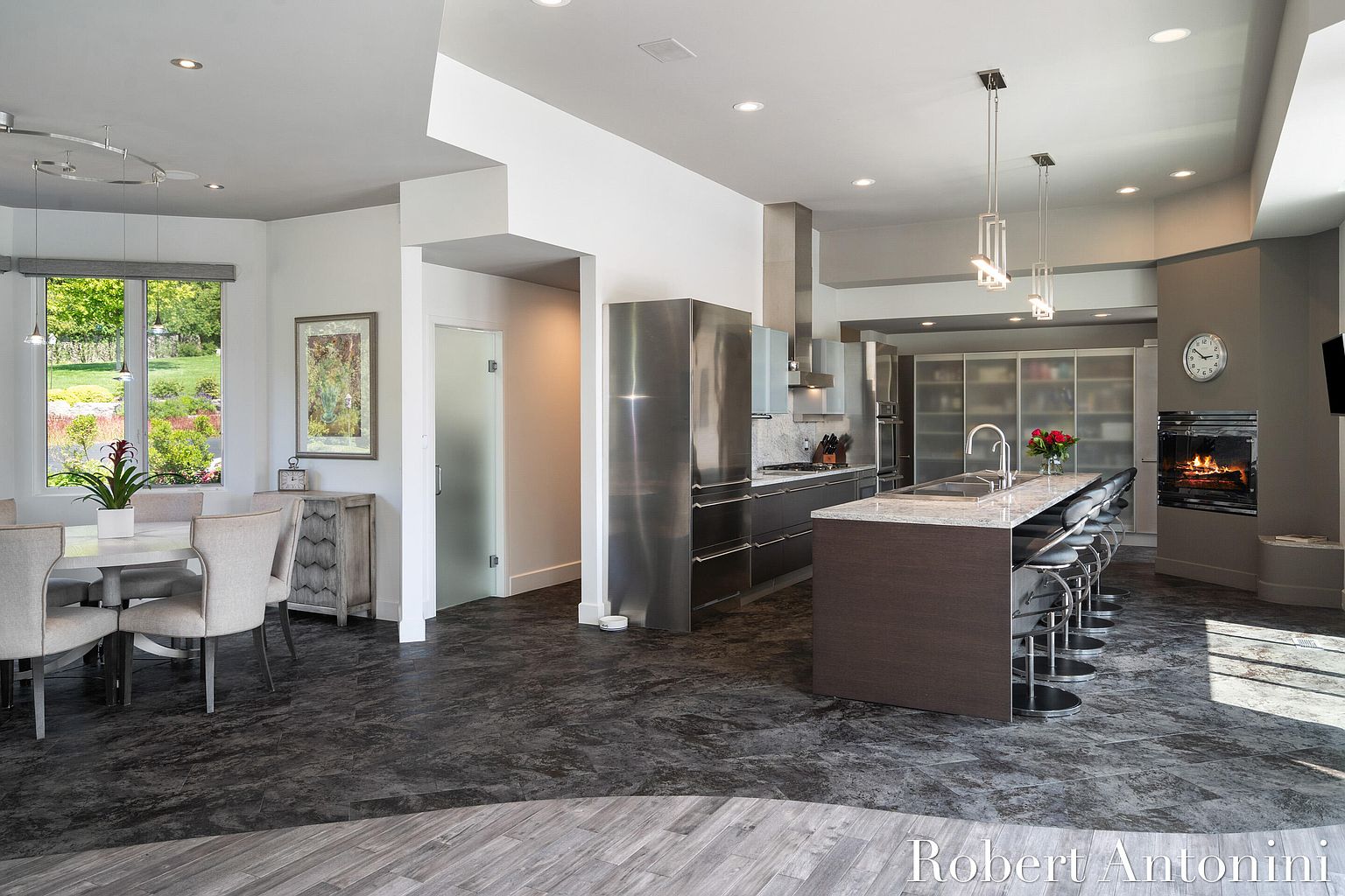 This is a spacious, modern kitchen with a large island featuring a marble countertop and seating for several people. Stainless steel appliances and sleek cabinetry contribute to the contemporary design. A fireplace adds a touch of warmth, and the open layout connects to a dining area with a view of the garden.