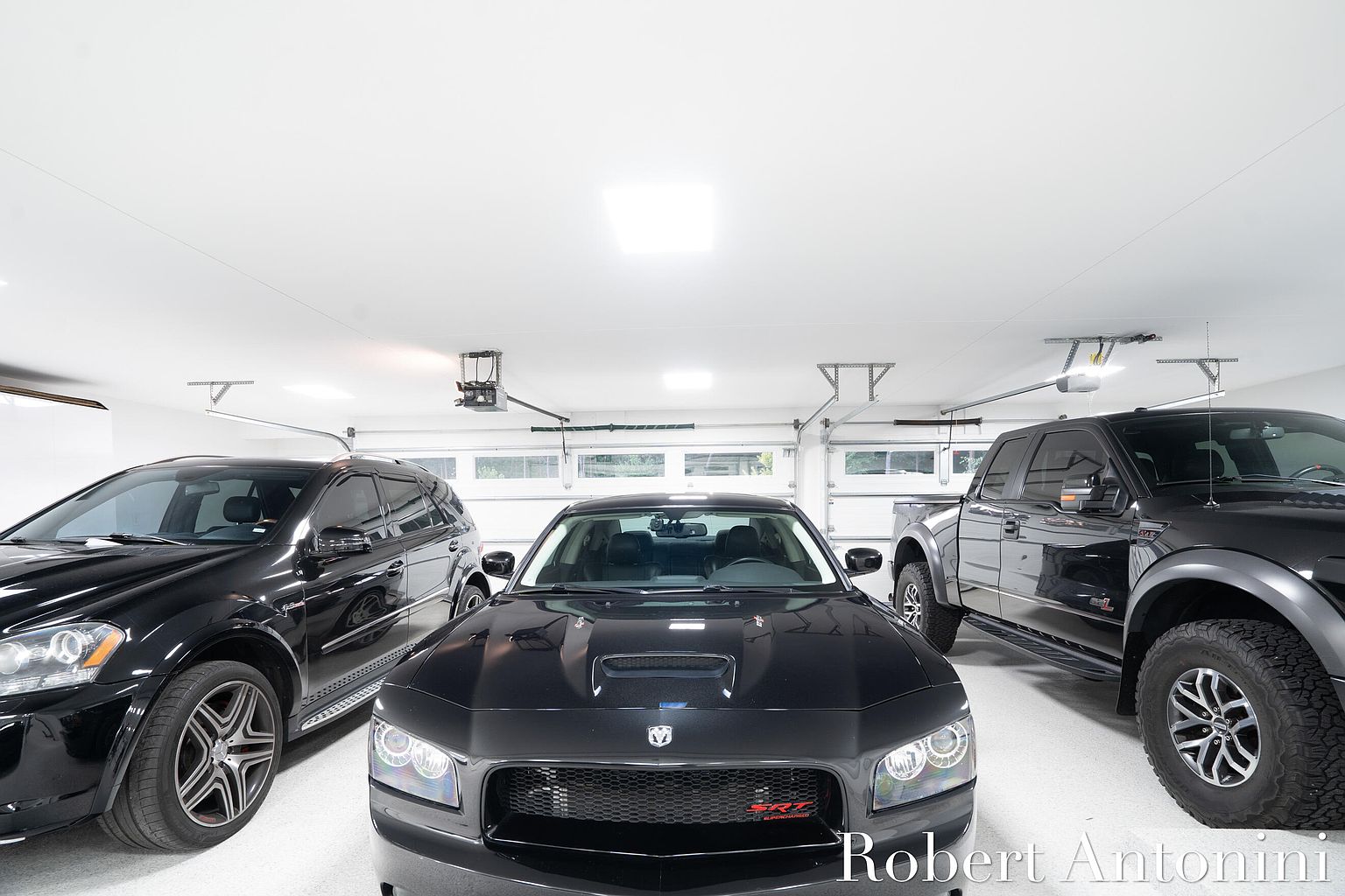 The image showcases a well-lit, organized garage featuring three black vehicles: a car in the center, an SUV on the left, and a truck on the right. The garage has a clean, white floor and walls, with visible garage door mechanisms and overhead lighting, creating a spacious and modern feel. The perspective is from the front, emphasizing the vehicles and the garage's overall layout.