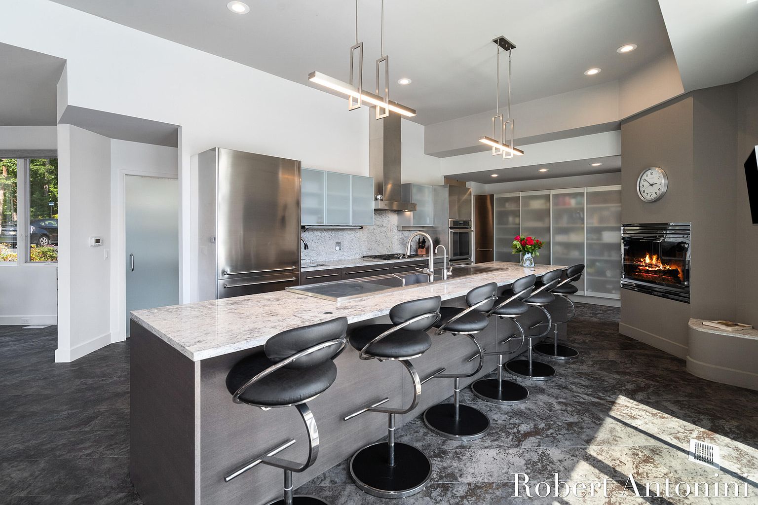 This is a modern kitchen with stainless steel appliances, including a large refrigerator and oven. The kitchen island features a light-colored countertop with seating for several people. A fireplace is visible on the right side of the image, adding a cozy element to the space.