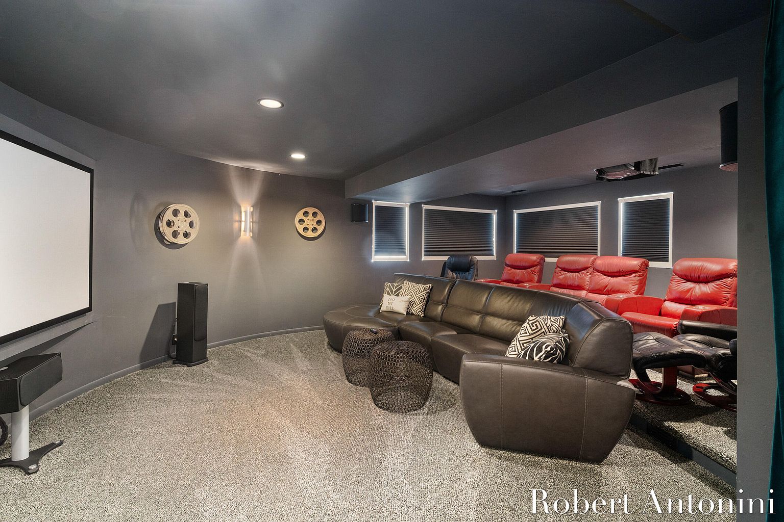 This is a home theater room in a basement, featuring a large projection screen, a curved leather sofa, and tiered seating with red leather recliners. The walls and ceiling are painted in a dark gray, creating a cinematic atmosphere, and the room is decorated with film reel accents. The perspective is from the front of the room, showcasing the seating arrangement and screen.