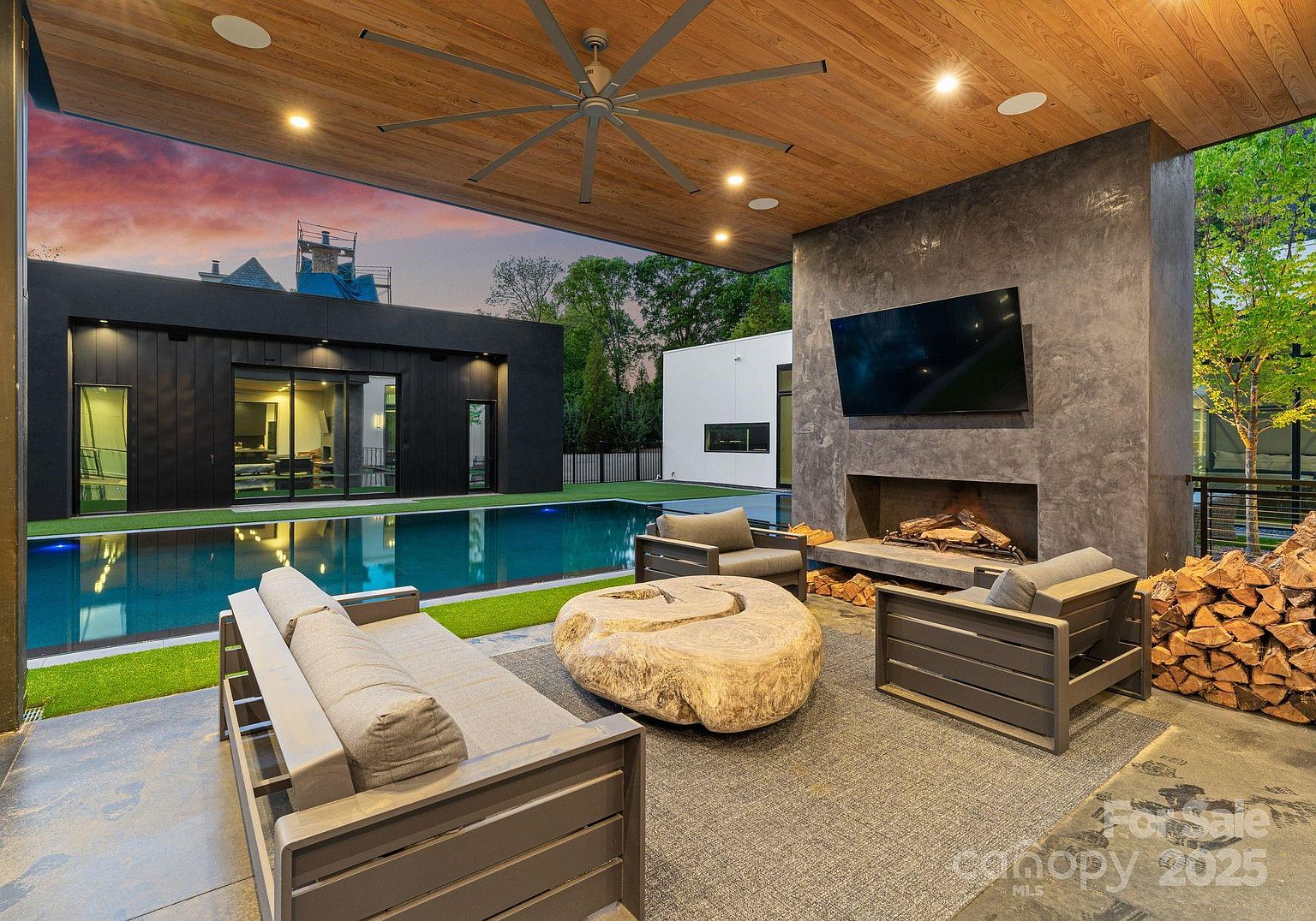 This modern covered patio features a large, natural-edge wood coffee table as a centerpiece, surrounded by contemporary outdoor seating. A tall, dark stone-clad chimney breast houses a fireplace and a mounted television, creating a sophisticated outdoor living space. The area overlooks a sleek swimming pool and a manicured lawn, with a warm wood-paneled ceiling and recessed lighting overhead.