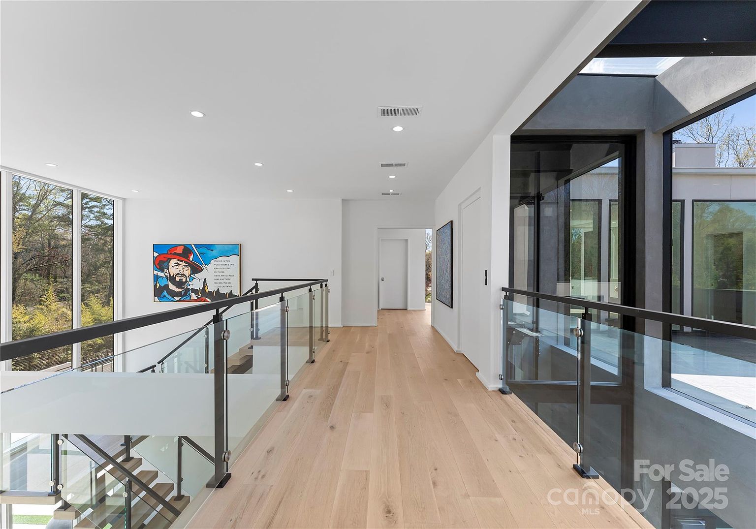 This bright, modern hallway features light wood flooring and a glass-paneled railing that overlooks a staircase. The space is characterized by clean white walls, recessed lighting, and large windows that provide an expansive view of the outdoors. A piece of colorful pop-art hangs on the wall, adding a vibrant focal point to the minimalist architectural design.