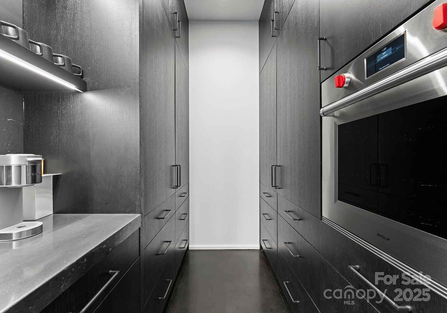 This sleek, modern butler's pantry or galley-style kitchen area features dark, textured cabinetry and a built-in Wolf wall oven. The space is defined by its narrow, symmetrical layout, with a countertop on the left and a clean, minimalist aesthetic throughout. The lighting is integrated under the upper cabinets, highlighting the dark surfaces and creating a sophisticated, high-end atmosphere.