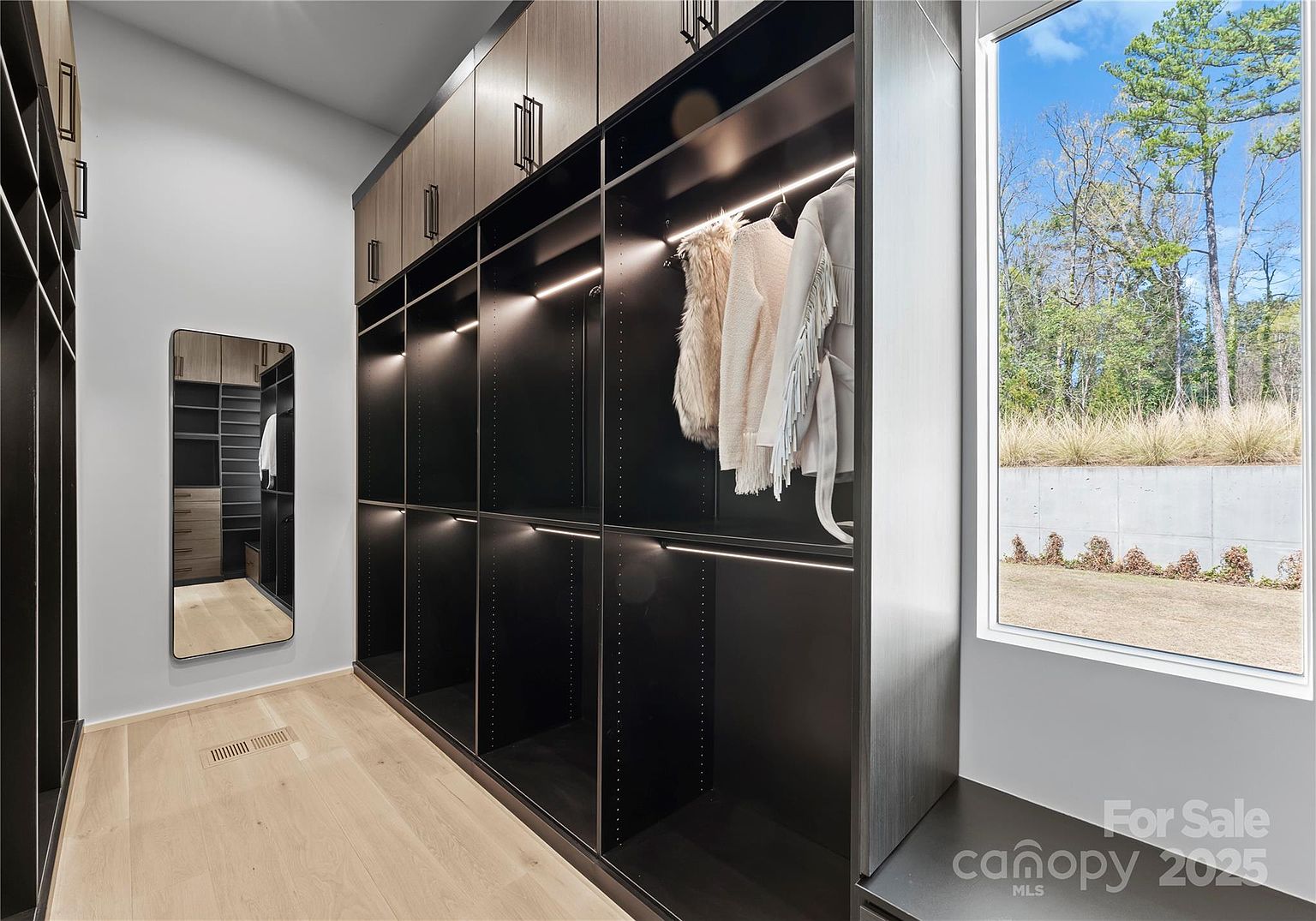 This modern walk-in closet features sleek, dark cabinetry with integrated LED lighting and a full-length mirror reflecting the organized storage space. A large window provides ample natural light and a view of the outdoor landscape, while light wood flooring adds warmth to the contemporary design. The clean lines and minimalist aesthetic create a sophisticated and functional dressing area.