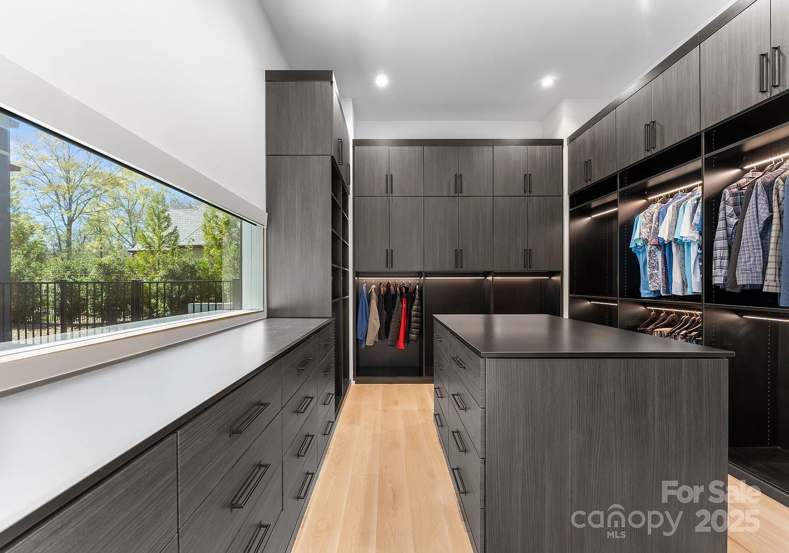 This spacious, modern walk-in closet features dark, wood-grain cabinetry with integrated lighting and sleek hardware. A central island provides additional storage and surface space, while a large window along the side wall offers a bright view of the outdoors. The room is finished with light wood flooring, creating a sophisticated and organized atmosphere.