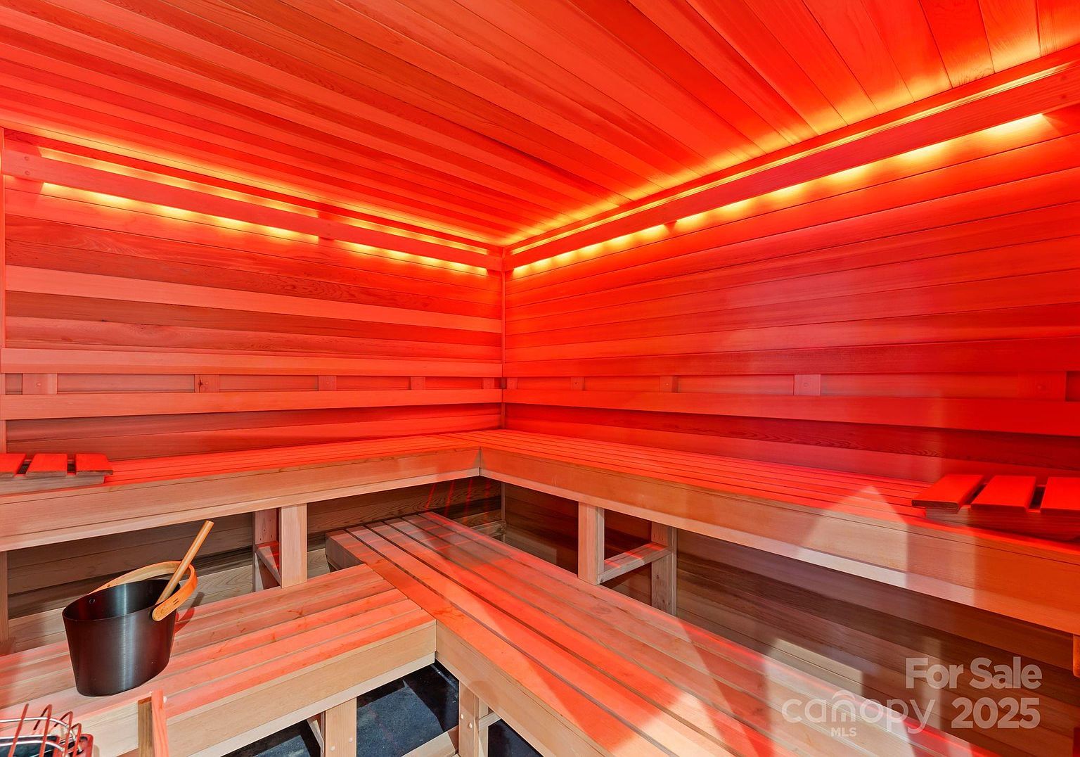 This image showcases a custom-built cedar sauna, characterized by its warm, aromatic wood paneling and tiered bench seating. The space is illuminated by integrated LED strip lighting that casts a soft, inviting glow across the horizontal wood slats. A metal bucket and ladle are positioned on the lower bench, emphasizing the authentic sauna experience.