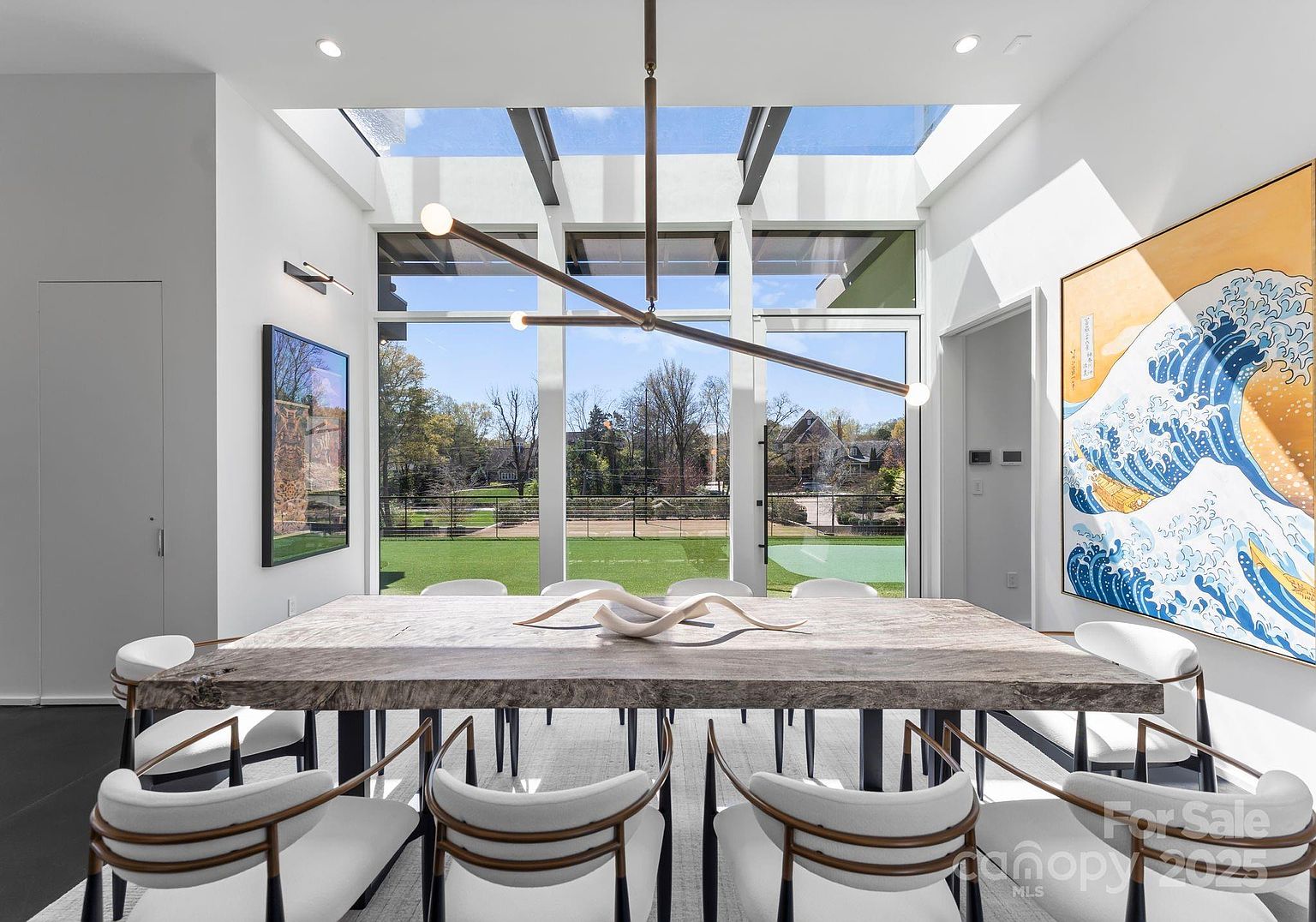 This modern dining room features a large, live-edge wood table surrounded by contemporary chairs with curved backs. The space is defined by expansive, floor-to-ceiling windows and a skylight that flood the room with natural light, offering a clear view of the outdoor landscape. A striking, multi-armed linear chandelier hangs above the table, while a large, vibrant mural of a stylized wave adds a bold artistic focal point to the side wall.