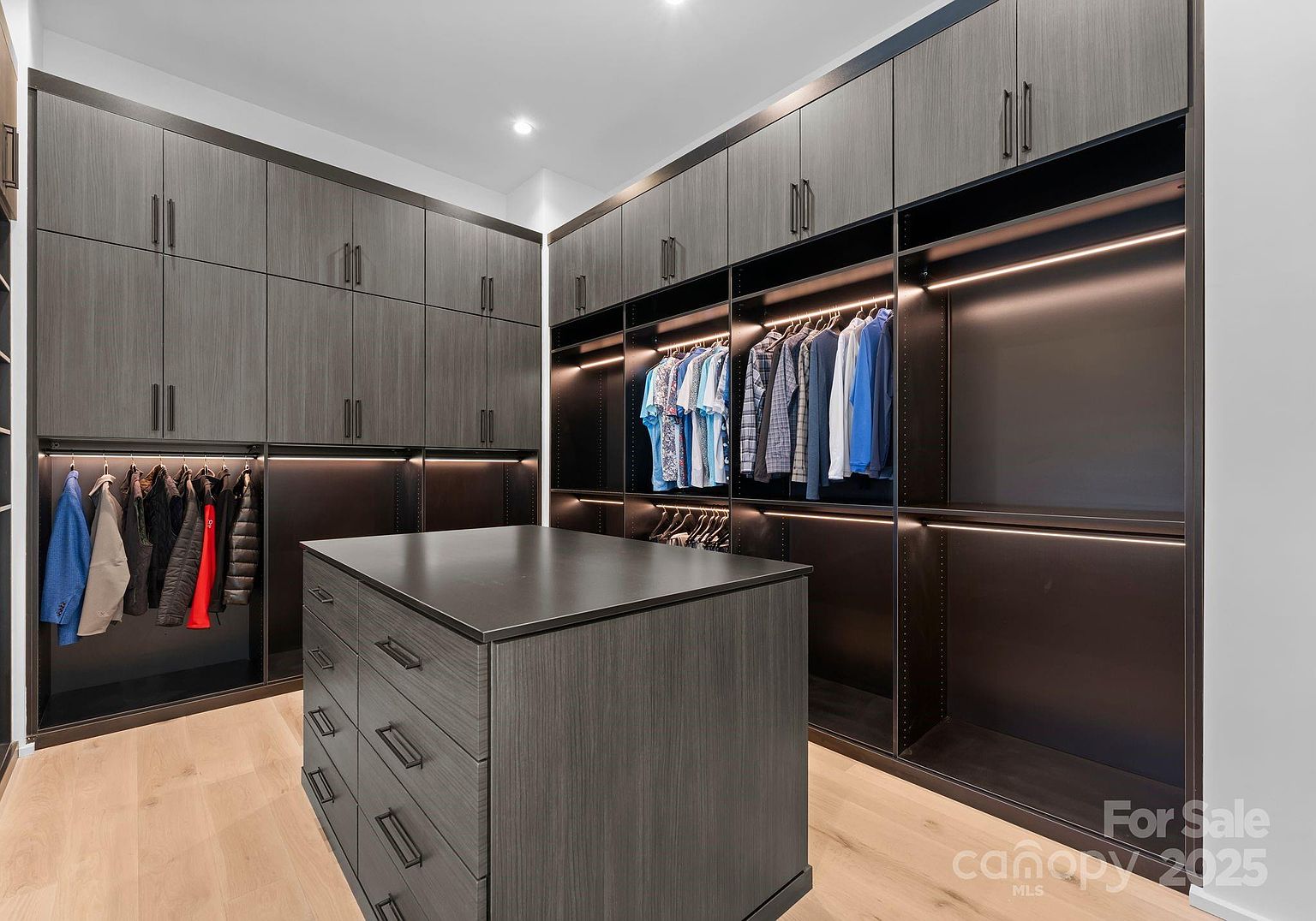 This spacious walk-in closet features dark, wood-grain cabinetry with integrated LED lighting and a central island with multiple drawers. The room is finished with light-toned wood flooring and provides ample hanging space for clothing, creating a sophisticated and organized storage environment. The perspective is from the entrance of the closet, showcasing the symmetrical layout and modern design.