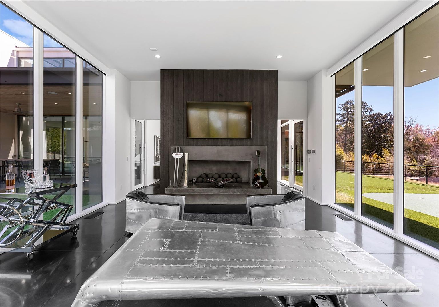 This modern living room features a striking dark wood-paneled chimney breast with a built-in television and a concrete fireplace hearth. The space is framed by expansive floor-to-ceiling windows that offer panoramic views of the outdoor landscape, while the room is furnished with unique metallic, industrial-style seating and a matching table. The polished dark flooring and minimalist aesthetic create a sophisticated, high-end atmosphere.