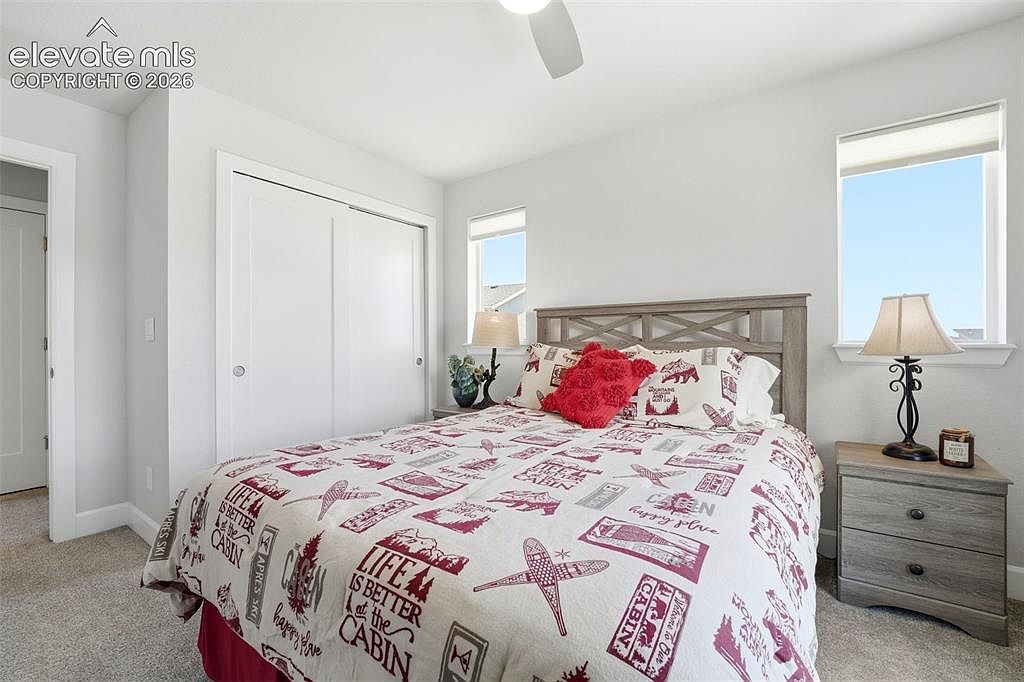 This cozy guest bedroom features a queen-sized bed with a rustic, cabin-themed comforter and a prominent red accent pillow. The room is framed by two windows that provide natural light, flanking a wooden headboard, and includes a matching bedside table with a lamp. The neutral wall color and carpeted flooring create a warm, inviting atmosphere suitable for a mountain or vacation home.