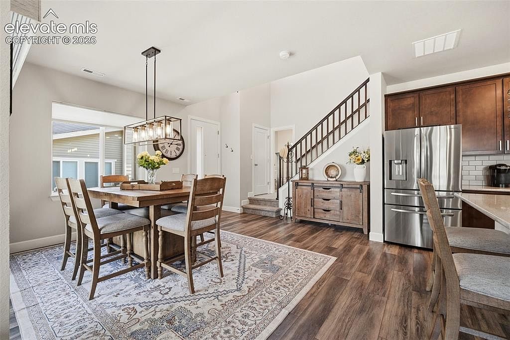 This open-concept dining area features a rustic wooden table surrounded by four chairs, set atop a large, patterned area rug. To the right, a wooden sideboard sits against the wall near a staircase with dark railings, while a stainless steel refrigerator and kitchen cabinetry are visible in the adjacent space. The room is illuminated by a modern rectangular pendant light fixture, creating a warm and inviting atmosphere.