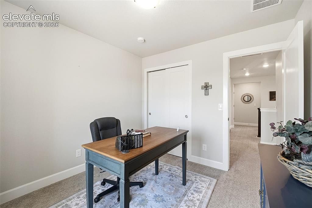 This home office features a central wooden desk paired with a black ergonomic chair, set atop a patterned area rug. The room is painted in a neutral light gray tone and includes a double-door closet, providing functional storage space. A doorway leads into a bright hallway, offering a glimpse into the rest of the home's interior layout.