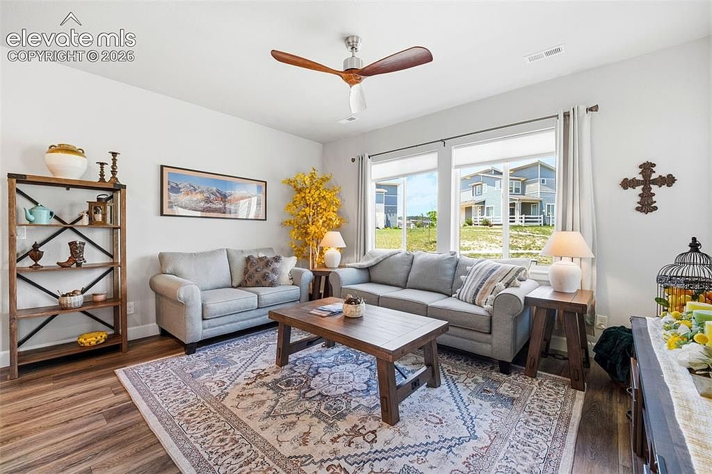 This inviting living room features a neutral color palette with two gray sofas arranged around a rustic wooden coffee table. A large, patterned area rug anchors the space, while a tall wooden shelving unit and a decorative yellow tree add character to the room. Large windows allow natural light to fill the area, providing a view of the neighborhood outside.