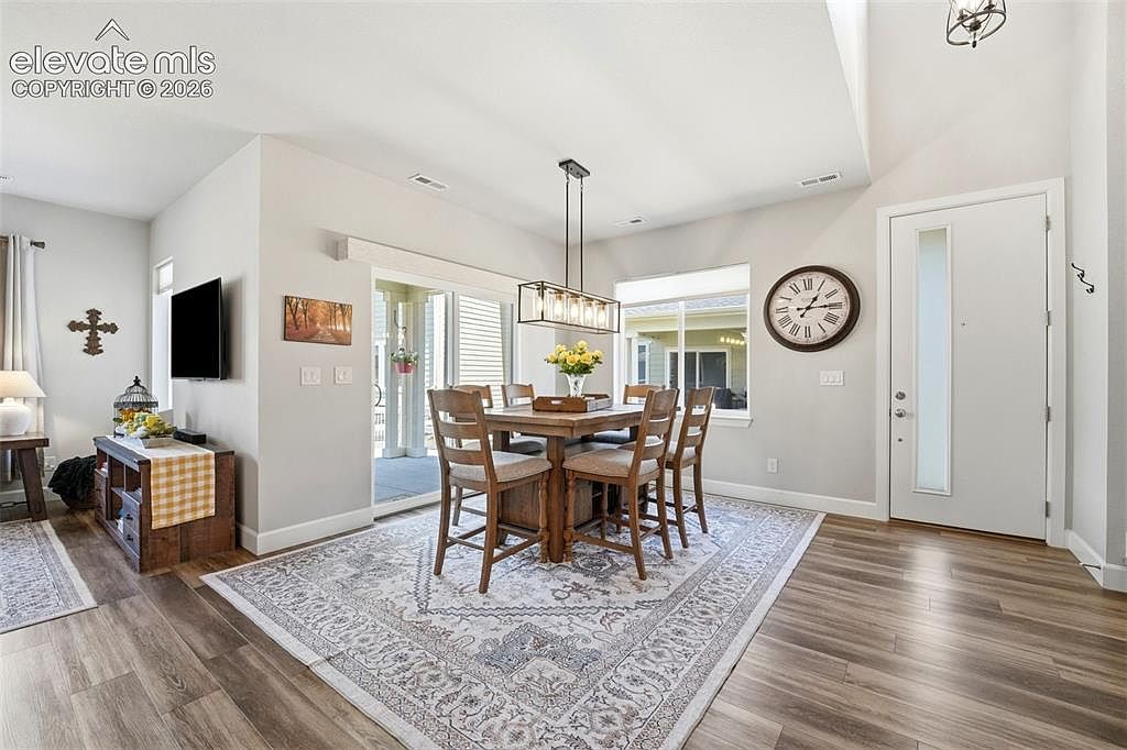 This bright and inviting dining area features a wooden table with four chairs set atop a large, patterned area rug. The space is illuminated by a modern rectangular pendant light fixture and includes a large wall clock, a television mounted on the wall, and a wooden console table. A sliding glass door provides a view of the outdoor patio, creating a seamless transition between indoor and outdoor living spaces.