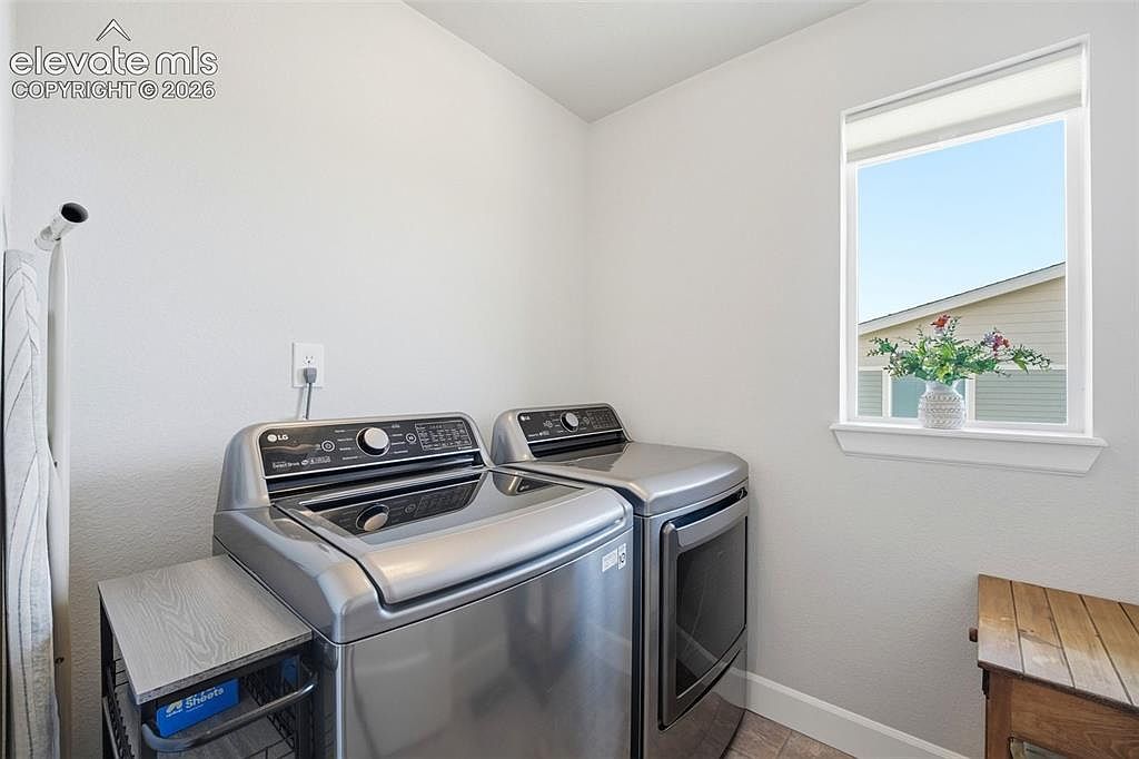 This functional laundry room features a pair of stainless steel LG washer and dryer units positioned against a neutral-toned wall. A window above the appliances allows natural light to brighten the space, while a small wooden side table and a wire storage rack provide convenient utility. The room has a clean, minimalist aesthetic, making it a practical and well-organized area for household chores.