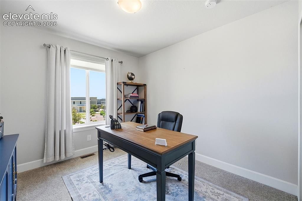This bright and functional home office features a central wooden desk paired with a black leather office chair, set atop a patterned area rug. A tall, open-shelving unit stands against the wall, providing storage for books and decor, while a large window with light-colored curtains allows for natural light and a view of the neighborhood. The room is finished with neutral walls and carpeted flooring, creating a clean and professional workspace.