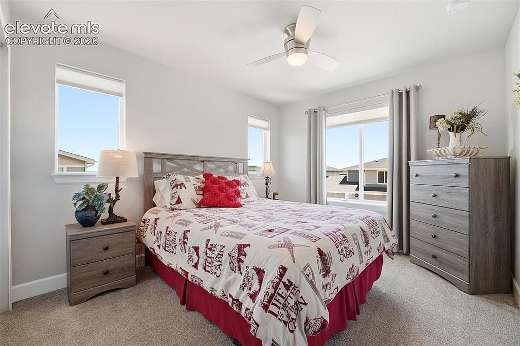 This bright and inviting bedroom features a queen-sized bed with a cabin-themed patterned comforter and a bold red accent pillow. The room is furnished with a matching light wood nightstand and a tall dresser, complemented by a ceiling fan and neutral wall tones. Large windows on two walls allow for ample natural light, creating a cozy and comfortable atmosphere.