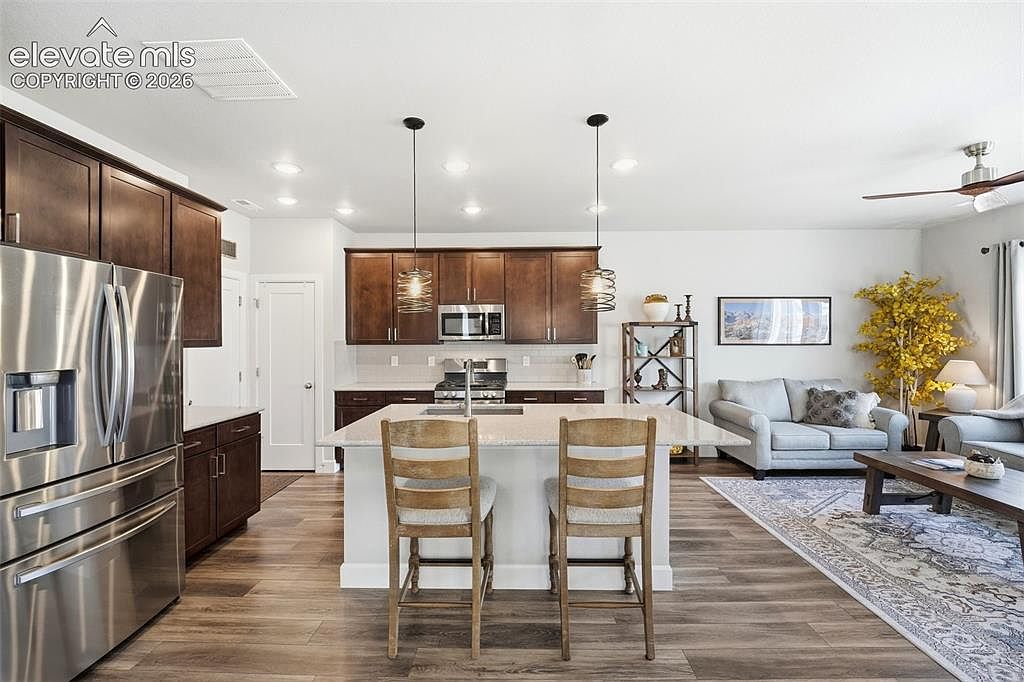 This open-concept kitchen features dark wood cabinetry, stainless steel appliances, and a central island with a light-toned countertop and two wooden bar stools. The space flows seamlessly into a living area, creating an inviting and functional layout with warm wood-look flooring throughout. Pendant lights hang above the island, providing focused illumination in this bright, modern home.