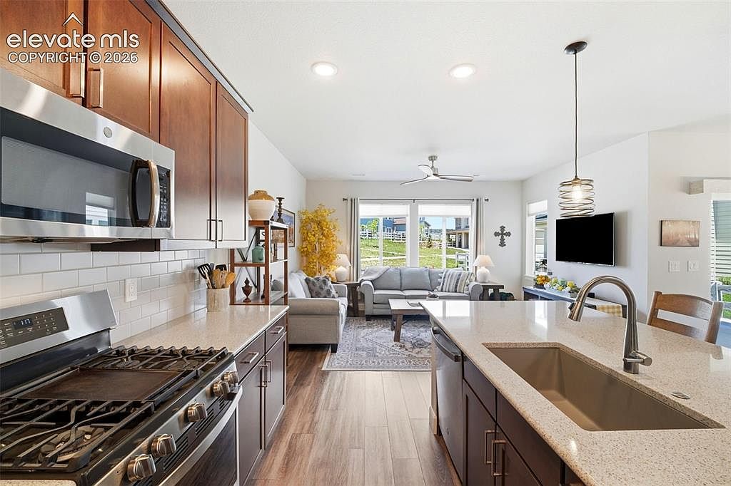 This open-concept kitchen features dark cabinetry, light-toned stone countertops, and stainless steel appliances, including a gas range and a built-in microwave. A large island with an undermount sink serves as a central focal point, seamlessly transitioning into the adjacent living area. The space is bright and airy, with wood-look flooring and a clear view through the living room to the outdoor scenery beyond.