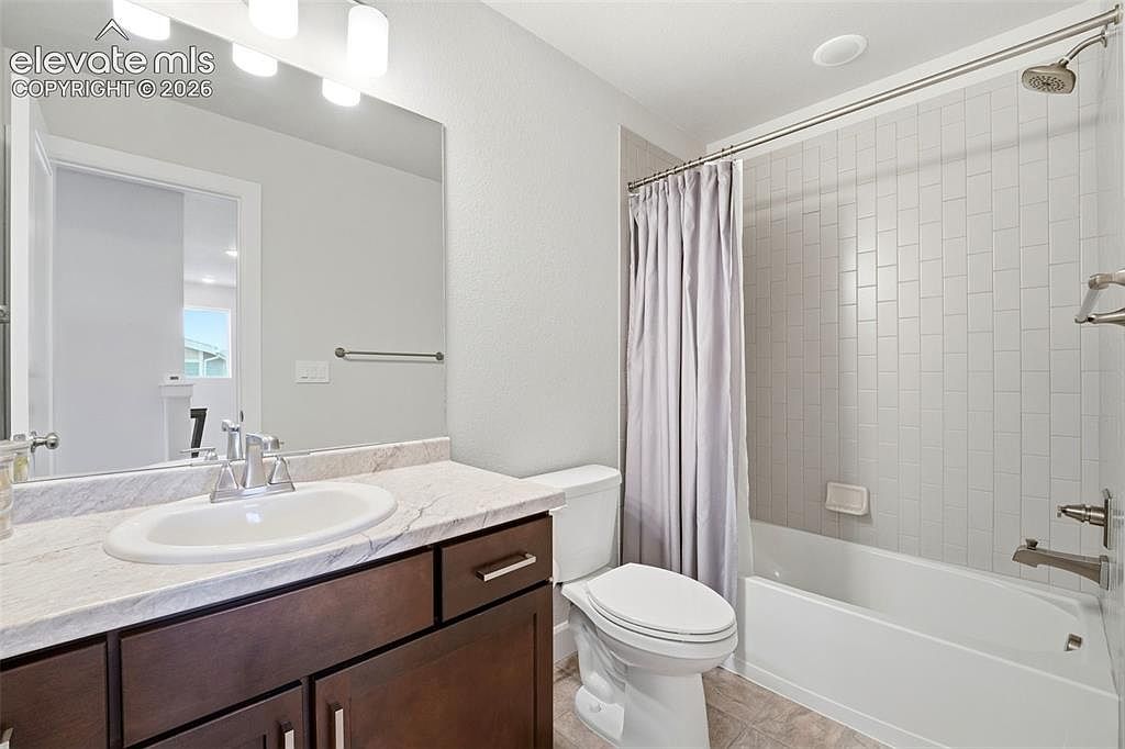 This clean and functional bathroom features a dark wood vanity topped with a light-toned stone counter and an integrated sink. A large, frameless mirror reflects the space, while a shower-tub combination is enclosed by a light gray curtain and finished with white subway-style wall tiles. The room is illuminated by a multi-bulb vanity light fixture and neutral wall paint, creating a bright and tidy atmosphere.
