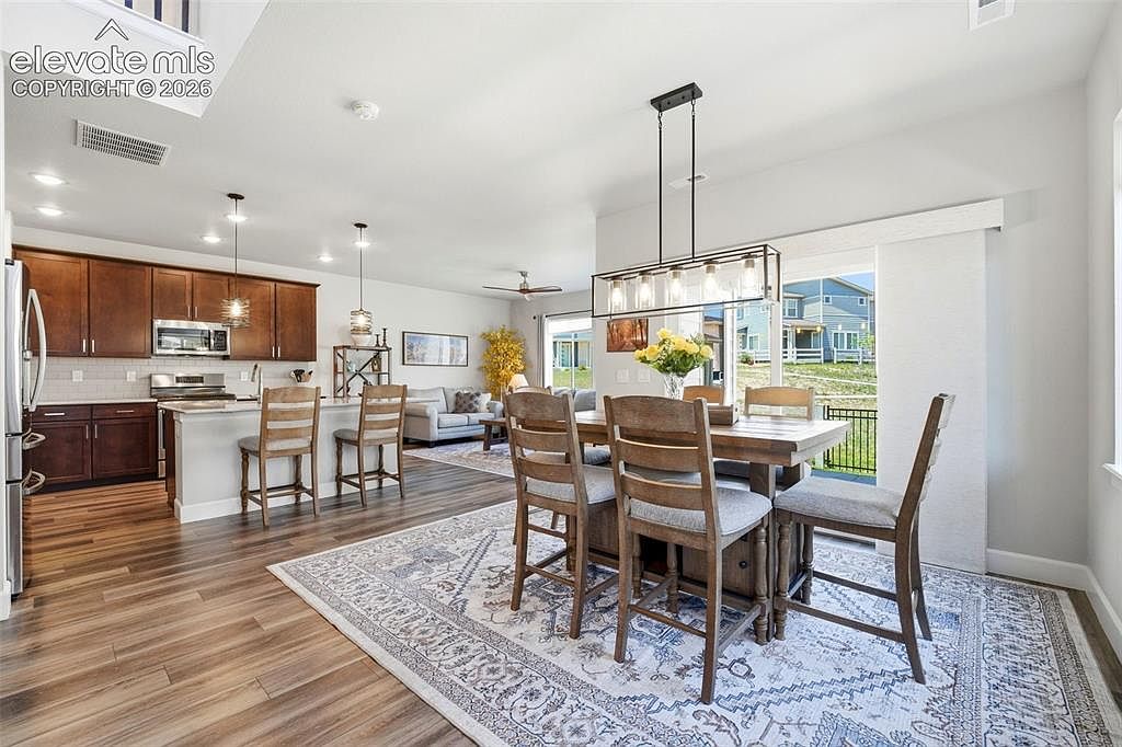 This open-concept space features a kitchen with dark wood cabinetry, stainless steel appliances, and a central island with bar seating. Adjacent to the kitchen is a dining area anchored by a large patterned rug and a modern linear pendant light fixture. The room flows seamlessly into a living area in the background, creating a bright and cohesive living space with wood-look flooring throughout.