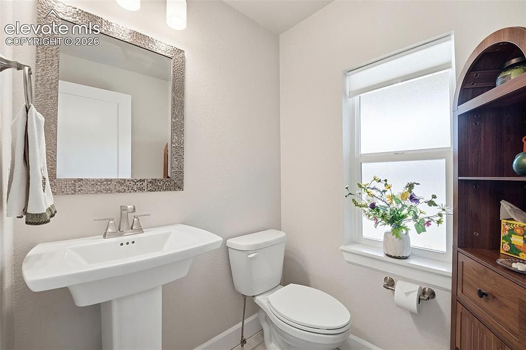 This bright and compact powder room features a white pedestal sink with a modern faucet and a textured, metallic-framed mirror above. A white toilet sits adjacent to a window that allows natural light to illuminate the space, which is decorated with a small vase of flowers on the sill. To the right, a tall, dark wood shelving unit provides storage, completing the functional and tidy aesthetic of the room.