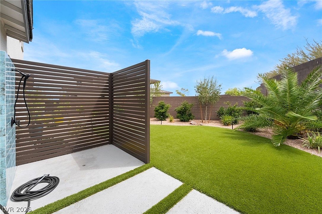 This outdoor space features a private, modern shower area enclosed by horizontal wood-slat privacy screens. The area includes a concrete patio pad transitioning into a well-manicured artificial turf lawn, bordered by desert-style landscaping and a tall masonry perimeter wall. The perspective captures a bright, sunny backyard setting, emphasizing a clean and low-maintenance lifestyle.