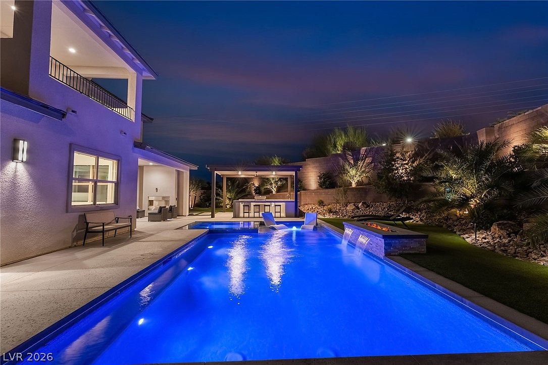 This evening shot captures a luxurious backyard oasis featuring a rectangular swimming pool with integrated water features and a raised spa. A covered outdoor kitchen area with bar seating sits in the background, complemented by manicured landscaping and ambient lighting that highlights the stone retaining wall. The perspective is from the poolside patio, showcasing a seamless transition between the home's exterior and the resort-style outdoor living space.