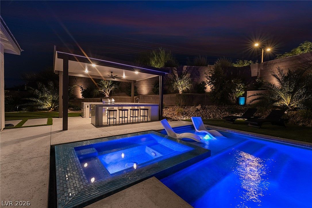 This nighttime view showcases a luxurious backyard oasis featuring a vibrant blue illuminated swimming pool and integrated spa. A covered outdoor kitchen area with bar seating provides a sophisticated space for entertaining, while strategically placed landscape lighting highlights the surrounding palm trees and stone retaining wall. The scene is captured from a low angle, emphasizing the inviting atmosphere of this high-end outdoor living space.