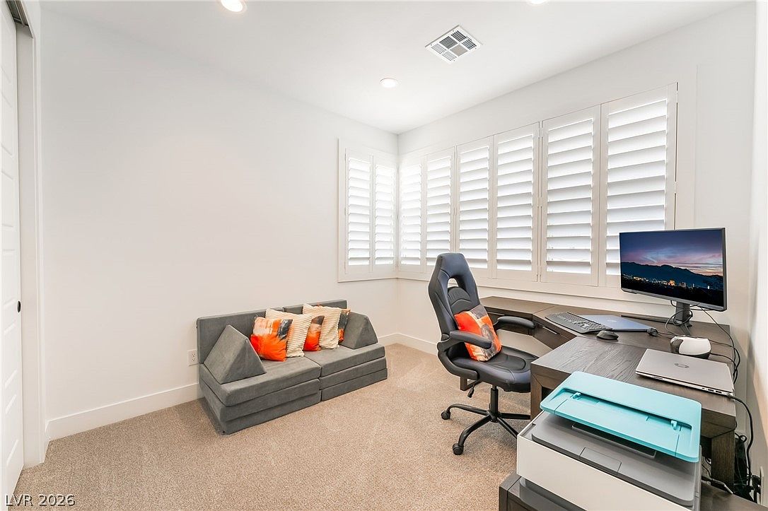 This bright and functional home office features a dark wood desk setup equipped with a computer monitor and a printer, paired with an ergonomic gaming-style chair. A modular, low-profile gray sofa with colorful accent pillows provides a comfortable seating area against the wall. The room is illuminated by recessed lighting and natural light streaming through large windows covered with white plantation shutters.