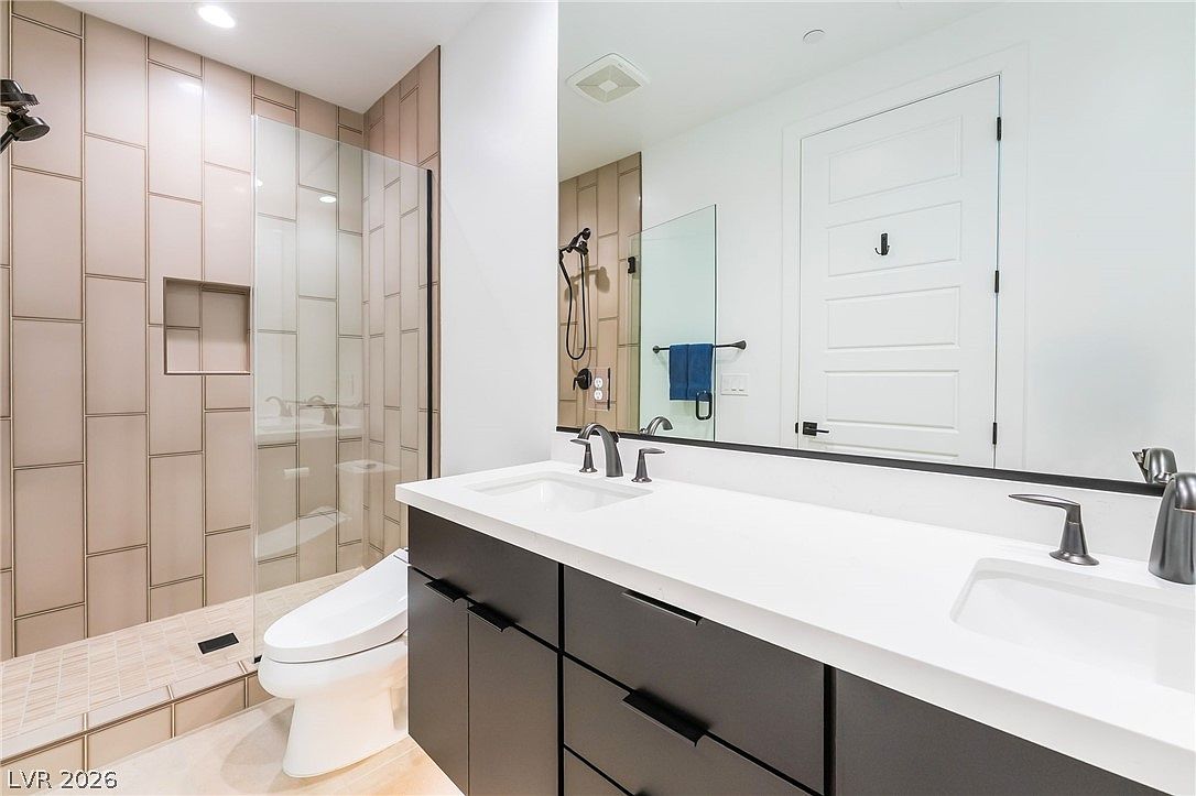 This modern bathroom features a double vanity with dark cabinetry, a white countertop, and a large mirror spanning the wall. To the left, a walk-in shower is enclosed by a glass panel and finished with light-toned, vertical rectangular tiles. The space is bright and contemporary, showcasing a clean, minimalist aesthetic with high-contrast finishes.