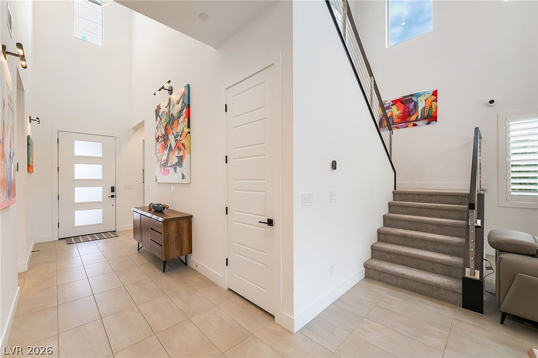 This bright and airy entryway features a high-ceiling foyer with light-toned tile flooring and a modern staircase with a metal railing. A wooden console table sits against the wall beneath a vibrant abstract painting, while a white door provides access to the home. The space is illuminated by natural light from tall windows, creating a welcoming and contemporary atmosphere.
